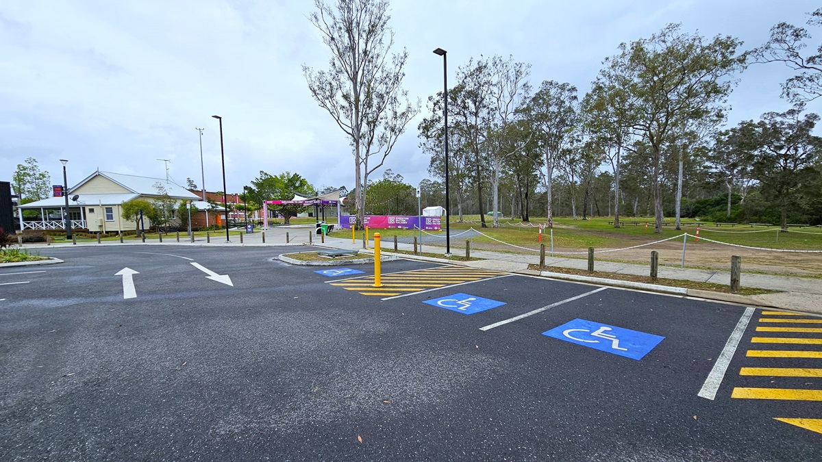 The parking area includes three accessible spaces on the right, marked with blue wheelchairs and yellow diagonal lines. A low rope fence, concrete bollards, and tall lamp posts line the edge, while a concrete plant box is positioned on the right. Trees and grass surround the area, leading to a white and brick building with colorful banners displaying a gradient shade of pink and blue. The parking area includes three accessible spaces on the right, marked with blue wheelchairs and yellow diagonal lines. A low rope fence, concrete bollards, and tall lamp posts line the edge, while a concrete plant box is positioned on the right. Trees and grass surround the area, leading to a white and brick building with colorful banners displaying a gradient shade of pink and blue.