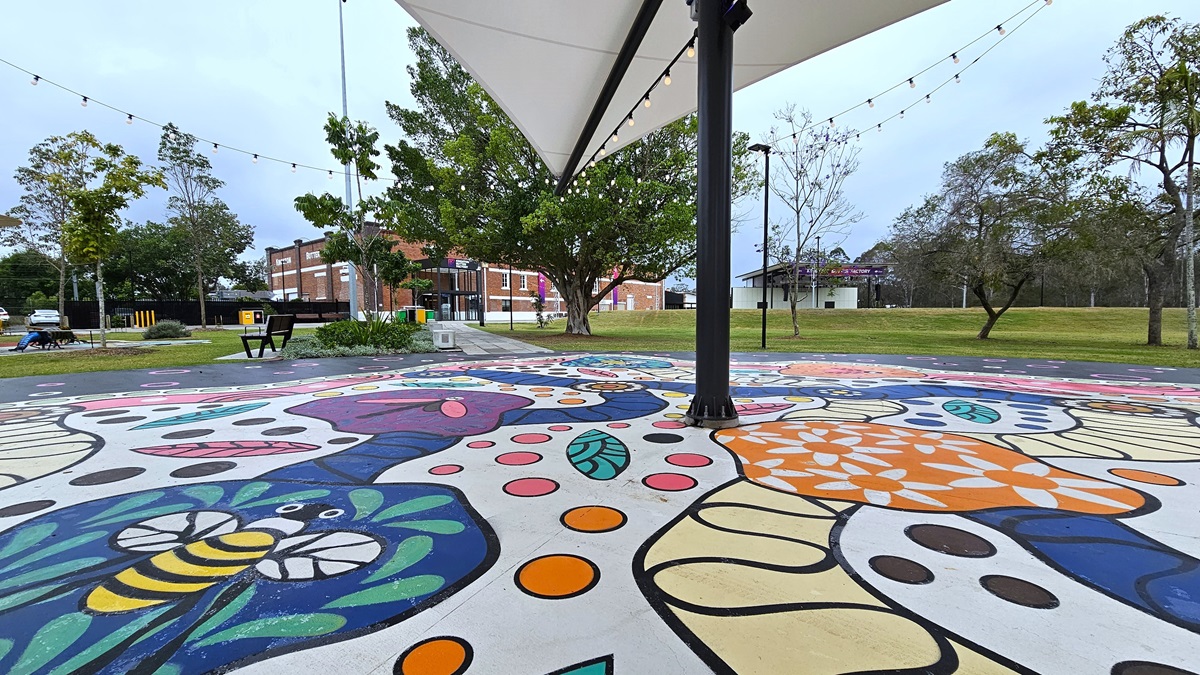In the center is a shade structure with light bulbs. Underneath, a colorful floor painting depicts imagery of bees, dragonflies, flowers, and leaves. On the left, a bench is positioned near a landscape plant box. Further ahead, a concrete pathway leads to a brick-red building. Tall trees and grass surround the area. In the center is a shade structure with light bulbs. Underneath, a colorful floor painting depicts imagery of bees, dragonflies, flowers, and leaves. On the left, a bench is positioned near a landscape plant box. Further ahead, a concrete pathway leads to a brick-red building. Tall trees and grass surround the area.