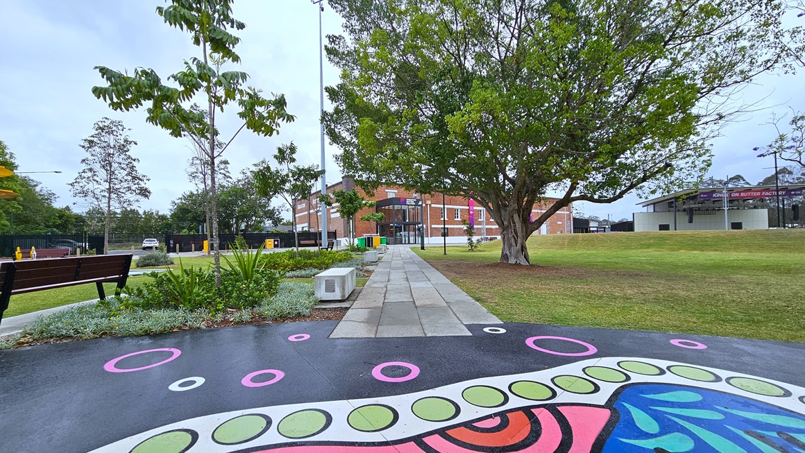 In the middle is a floor with colorful prints, connected to a concrete path leading to the brick-red building. On the left, a sitting bench is surrounded by ornamental plants and trees. Stone structures with inscriptions are lined up on the left. To the right, a tall tree with robust branches stands, and further right is a stage. Green grass surrounds the area on both sides. In the middle is a floor with colorful prints, connected to a concrete path leading to the brick-red building. On the left, a sitting bench is surrounded by ornamental plants and trees. Stone structures with inscriptions are lined up on the left. To the right, a tall tree with robust branches stands, and further right is a stage. Green grass surrounds the area on both sides.