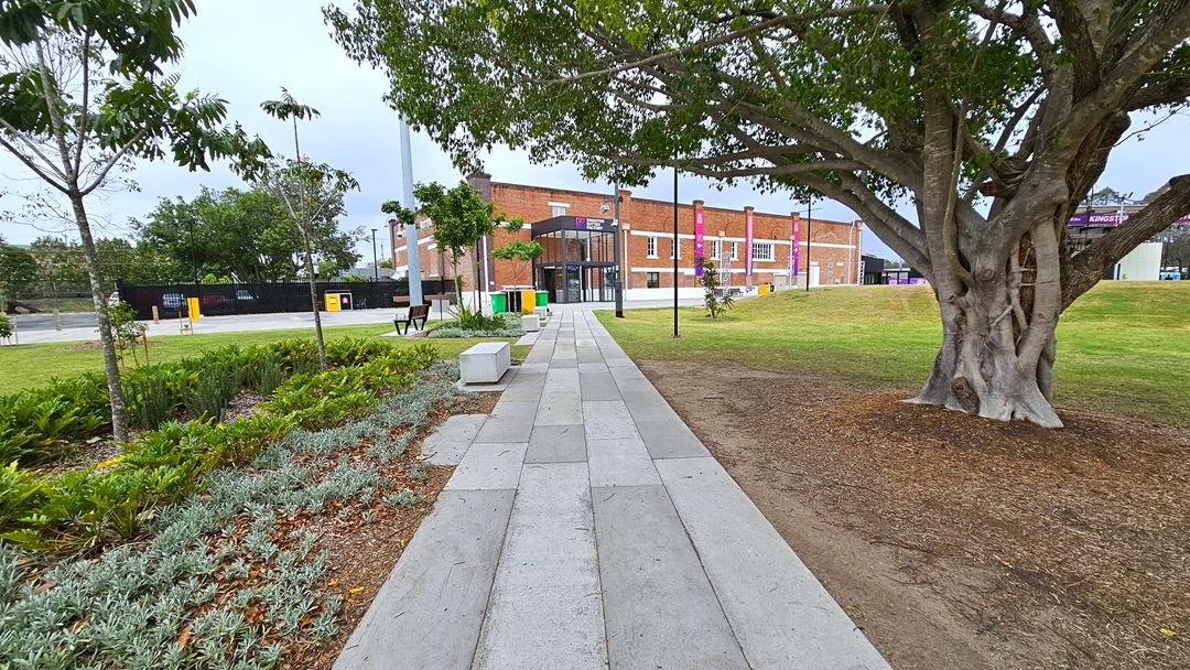In the middle is a concrete path leading to the red brick building adorned with gradient banners. On the left, there's a landscape of ornamental plants and trees, with a bench for sitting further ahead. Stone structures with inscriptions are lined up on the left. To the right, a tall tree with robust branches stands, and further right is a stage. Green grass surrounds the area on both sides. In the middle is a concrete path leading to the red brick building adorned with gradient banners. On the left, there's a landscape of ornamental plants and trees, with a bench for sitting further ahead. Stone structures with inscriptions are lined up on the left. To the right, a tall tree with robust branches stands, and further right is a stage. Green grass surrounds the area on both sides.