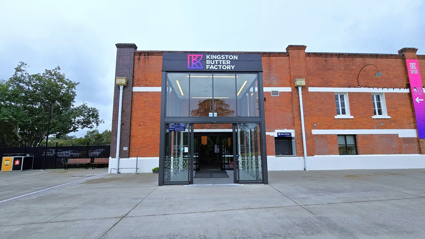 In the middle stands an industrial building made of red bricks with glass windows. Its front is adorned with a gradient blue and pink banner, vertically hung on the right side along with light fixtures. The entrance, a double modern glass and steel structure, features facility signage atop the glass door. On the left, a black steel gate and rubbish bins are present. Trees are also within the fenced area. In the middle stands an industrial building made of red bricks with glass windows. Its front is adorned with a gradient blue and pink banner, vertically hung on the right side along with light fixtures. The entrance, a double modern glass and steel structure, features facility signage atop the glass door. On the left, a black steel gate and rubbish bins are present. Trees are also within the fenced area.