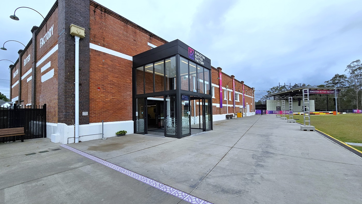In the middle is an industrial building constructed from red bricks, featuring glass windows. The front is decorated with a gradient blue and pink banner, vertically suspended on the right alongside light fixtures. The entrance, a double modern glass and steel structure, displays facility signage above the glass door. On the left, there's a black steel fence with a bench for seating. To the right, a stage is adorned with gradient banners, and nearby, there's a grassy area with steel structures. In the middle is an industrial building constructed from red bricks, featuring glass windows. The front is decorated with a gradient blue and pink banner, vertically suspended on the right alongside light fixtures. The entrance, a double modern glass and steel structure, displays facility signage above the glass door. On the left, there's a black steel fence with a bench for seating. To the right, a stage is adorned with gradient banners, and nearby, there's a grassy area with steel structures.