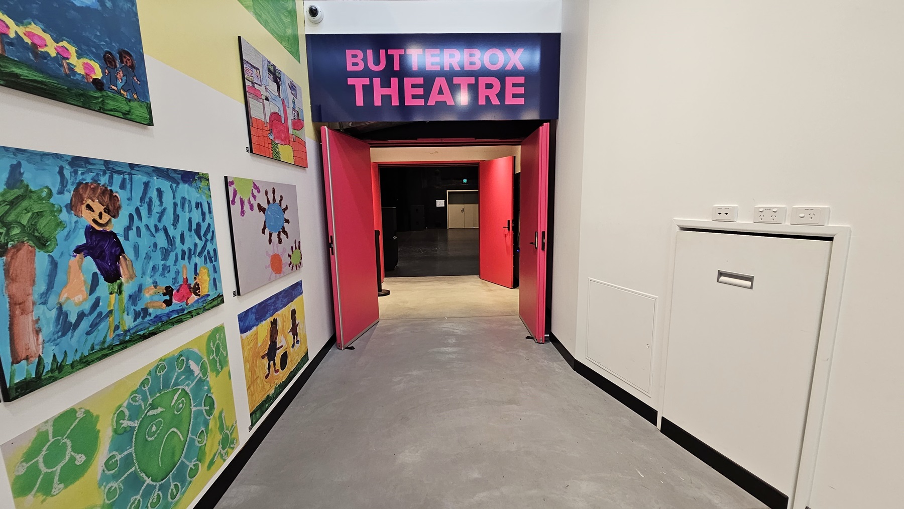 To the left, a wall features adorned art paintings. In the middle, a bright pink door opens outward, accompanied by an overhead sign displaying "Butterbox Theatre" in pink font. On the right, a white concierge table is placed adjacent to the white wall. The floor is finished with polished concrete. To the left, a wall features adorned art paintings. In the middle, a bright pink door opens outward, accompanied by an overhead sign displaying "Butterbox Theatre" in pink font. On the right, a white concierge table is placed adjacent to the white wall. The floor is finished with polished concrete.