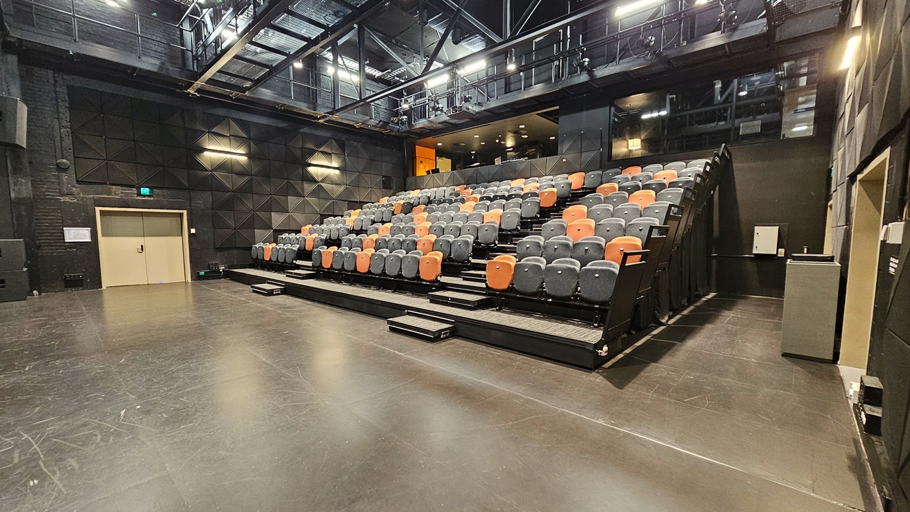 On the right, tiered seating features black and orange chairs separated by an aisle. To the left, a drinking fountain is present. An elevated area is equipped with chairs. The ceiling adopts an industrial look with exposed beams and fluorescent lights. The area has dimmed lighting, black wall tiles, complemented by grey tiles. A door on the left is marked with an Emergency Exit sign on top. On the right, tiered seating features black and orange chairs separated by an aisle. To the left, a drinking fountain is present. An elevated area is equipped with chairs. The ceiling adopts an industrial look with exposed beams and fluorescent lights. The area has dimmed lighting, black wall tiles, complemented by grey tiles. A door on the left is marked with an Emergency Exit sign on top.