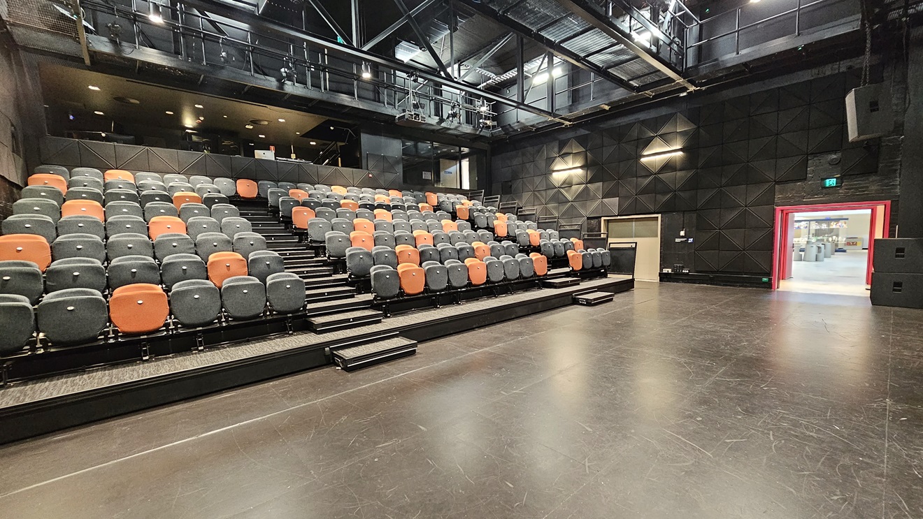 In the middle, tiered seating features black and orange chairs separated by an aisle. An elevated area is equipped with chairs. The ceiling adopts an industrial look with exposed beams and fluorescent lights. The area has dimmed lighting, and black wall tiles complemented by grey tiles. A bright pink door on the right has an Emergency Exit sign on top, and another white door to its left. The wall is soundproofed with black tiles. In the middle, tiered seating features black and orange chairs separated by an aisle. An elevated area is equipped with chairs. The ceiling adopts an industrial look with exposed beams and fluorescent lights. The area has dimmed lighting, and black wall tiles complemented by grey tiles. A bright pink door on the right has an Emergency Exit sign on top, and another white door to its left. The wall is soundproofed with black tiles.