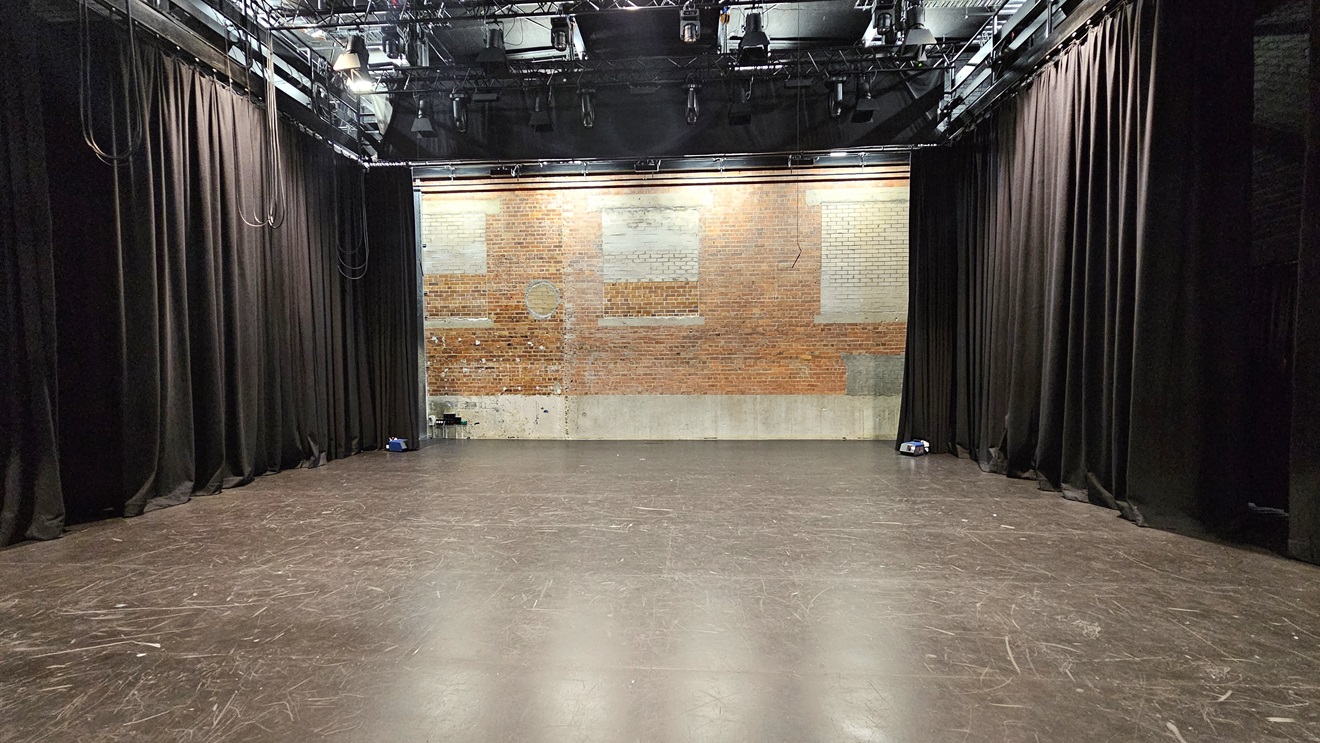 In the middle, there's a stage with a red brick wall backdrop. The stage area contains substantial theatre production equipment, featuring boom lighting and large black drop curtains against the left and right brick walls. The floor is composed of grey tiles. In the middle, there's a stage with a red brick wall backdrop. The stage area contains substantial theatre production equipment, featuring boom lighting and large black drop curtains against the left and right brick walls. The floor is composed of grey tiles.