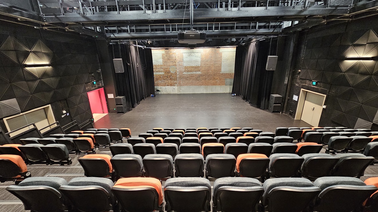 The tiered seating consists of orange and black chairs, with an aisle on the left dividing them. In front is the stage, featuring black drop curtains. A pink door is on the left, and a white door is on the right. The ceiling is adorned with industrial metal beams. Rubber tiles are affixed to the walls on both sides. The tiered seating consists of orange and black chairs, with an aisle on the left dividing them. In front is the stage, featuring black drop curtains. A pink door is on the left, and a white door is on the right. The ceiling is adorned with industrial metal beams. Rubber tiles are affixed to the walls on both sides.