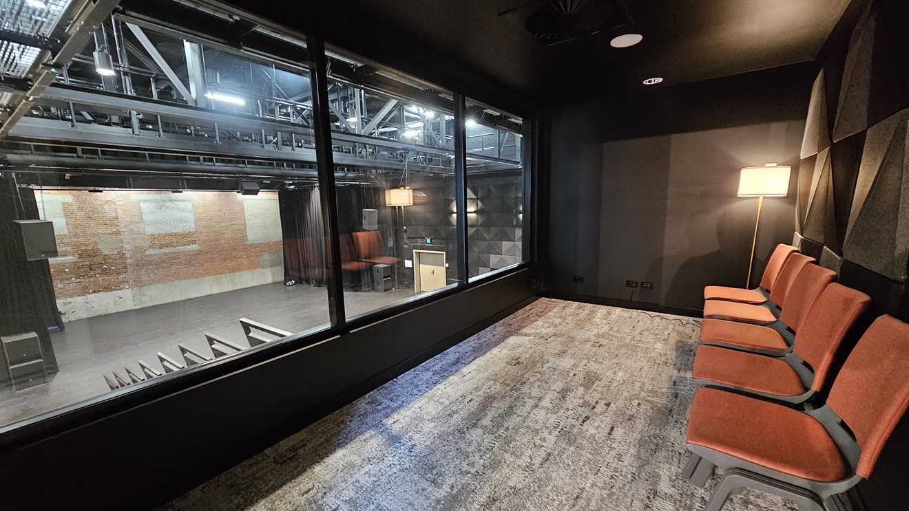 This room has orange chairs arranged on one side, enclosed by glass, overlooking the stage. The wall is surrounded by black rubber tiles, and the wall itself is black. The room is partially dim. In front is the stage with black drop curtains, and the ceiling features industrial metal beams. Rubber tiles are affixed to both sides of the walls. This room has orange chairs arranged on one side, enclosed by glass, overlooking the stage. The wall is surrounded by black rubber tiles, and the wall itself is black. The room is partially dim. In front is the stage with black drop curtains, and the ceiling features industrial metal beams. Rubber tiles are affixed to both sides of the walls.