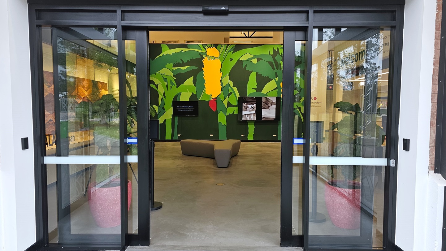 The entrance is an automatic glass sliding door with a white sticker lining. Inside, a Y-shaped cushioned chair is in the middle. A green wall featuring banana trees with fruit is present. Two flat screens are mounted. To the right is the reception. A pot of ornamental plants is positioned near the door entrance. The entrance is an automatic glass sliding door with a white sticker lining. Inside, a Y-shaped cushioned chair is in the middle. A green wall featuring banana trees with fruit is present. Two flat screens are mounted. To the right is the reception. A pot of ornamental plants is positioned near the door entrance.