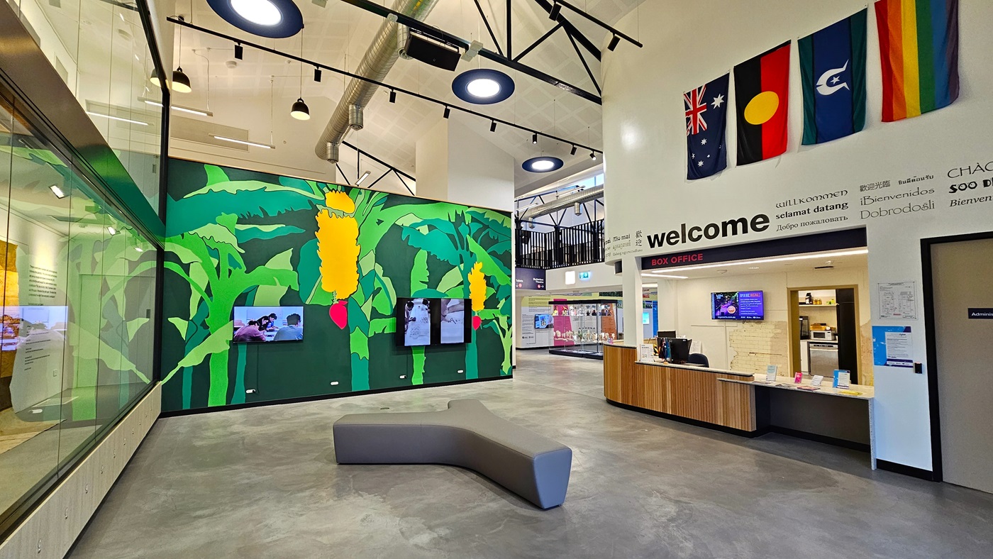 In the middle is a Y-shaped cushioned chair. A green wall featuring banana trees with fruit is present. To the right is the reception, adorned with a wooden desk displaying various posters and flags. Flat-screen TVs are also mounted on the middle wall and at the reception. Banners are affixed to the right wall. To the left is a floor-to-ceiling glass wall. The ceiling is adorned with circular light fixtures. On the far right, a section of the mezzanine is visible. In the middle is a Y-shaped cushioned chair. A green wall featuring banana trees with fruit is present. To the right is the reception, adorned with a wooden desk displaying various posters and flags. Flat-screen TVs are also mounted on the middle wall and at the reception. Banners are affixed to the right wall. To the left is a floor-to-ceiling glass wall. The ceiling is adorned with circular light fixtures. On the far right, a section of the mezzanine is visible.