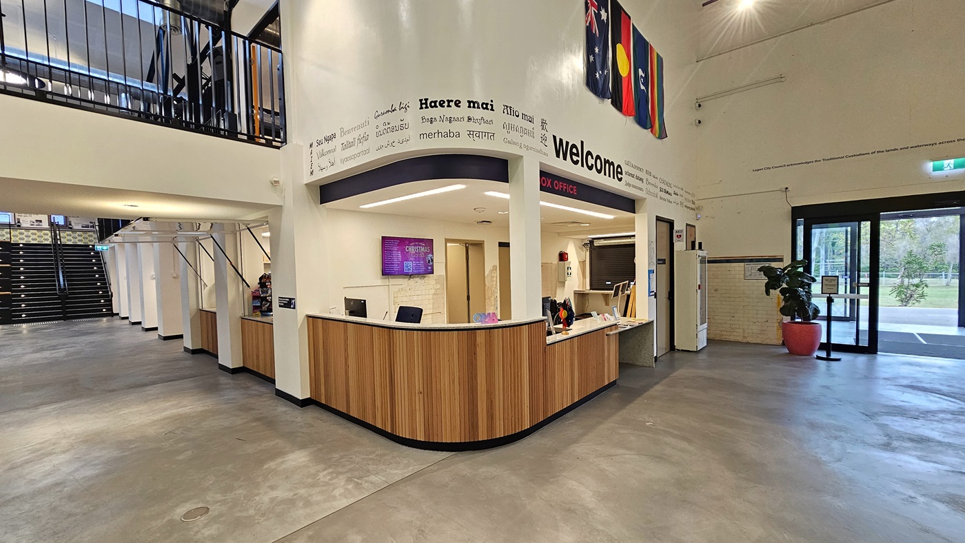 On the right is the reception, featuring a wooden desk with various flags. Flat-screen TVs are mounted on the middle wall of the reception. Banners and posters are affixed to the right wall. To the left is a staircase leading to the mezzanine. A section of the mezzanine is also visible. To the right is the door entrance, with a pot of ornamental plants positioned near it. On the right is the reception, featuring a wooden desk with various flags. Flat-screen TVs are mounted on the middle wall of the reception. Banners and posters are affixed to the right wall. To the left is a staircase leading to the mezzanine. A section of the mezzanine is also visible. To the right is the door entrance, with a pot of ornamental plants positioned near it.