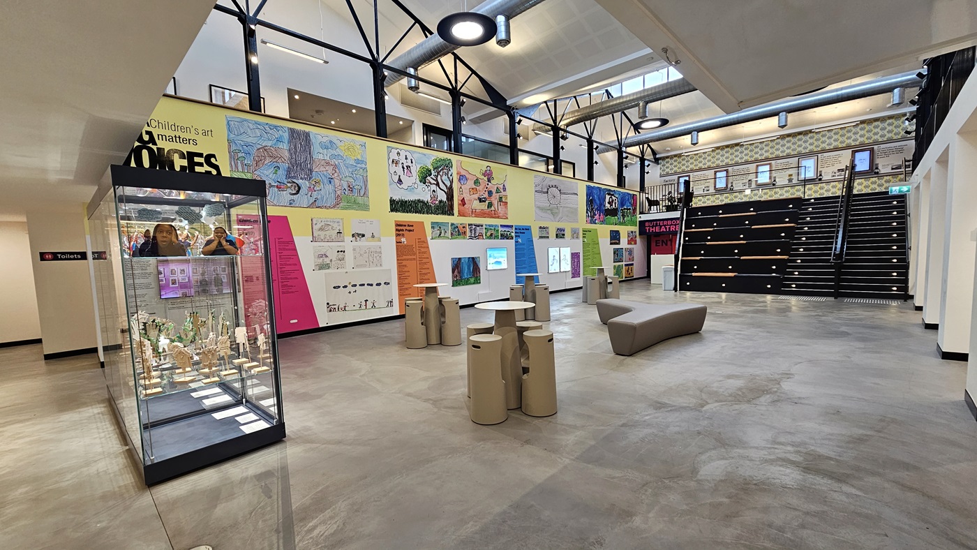 In the centre is the reception, alongside a small café. The desks in both the reception and café are designed with a wooden timber motif. Flat-screen TVs are mounted on the walls. To the left is an area leading to an open space with seating. The mezzanine level is also visible. The area is well-lit with light fixtures and fluorescent lights. In the centre is the reception, alongside a small café. The desks in both the reception and café are designed with a wooden timber motif. Flat-screen TVs are mounted on the walls. To the left is an area leading to an open space with seating. The mezzanine level is also visible. The area is well-lit with light fixtures and fluorescent lights.