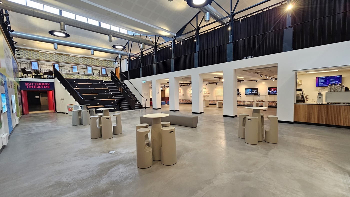 On the left, the wall displays various posters, paintings, and art materials. Bright pink entrance doors are situated on the left. In the middle, there are Y-shaped cream-cushioned chairs along with uniquely designed stools and tables. A stair leading to the mezzanine and tiered seating is also positioned in the middle. The ceiling features an open space with circular lights and large air-conditioning vents. The cafe is on the right, and further ahead is a spacious seating area. On the left, the wall displays various posters, paintings, and art materials. Bright pink entrance doors are situated on the left. In the middle, there are Y-shaped cream-cushioned chairs along with uniquely designed stools and tables. A stair leading to the mezzanine and tiered seating is also positioned in the middle. The ceiling features an open space with circular lights and large air-conditioning vents. The cafe is on the right, and further ahead is a spacious seating area.