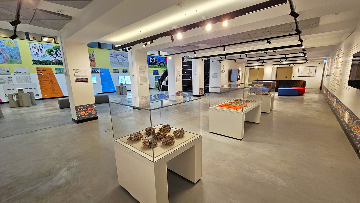 On the right is a spacious area with cushioned seating in the corner. Three tables with enclosed glass displays are present. The right wall showcases meticulously painted wall art, with large flat-screen TVs mounted on it. The ceiling is white, adorned with light fixtures and ceiling art decoration. The floor area is made of polished concrete. The right wall is adorned with art posters and paintings, and there are uniquely designed stools and tables for seating. On the right is a spacious area with cushioned seating in the corner. Three tables with enclosed glass displays are present. The right wall showcases meticulously painted wall art, with large flat-screen TVs mounted on it. The ceiling is white, adorned with light fixtures and ceiling art decoration. The floor area is made of polished concrete. The right wall is adorned with art posters and paintings, and there are uniquely designed stools and tables for seating.