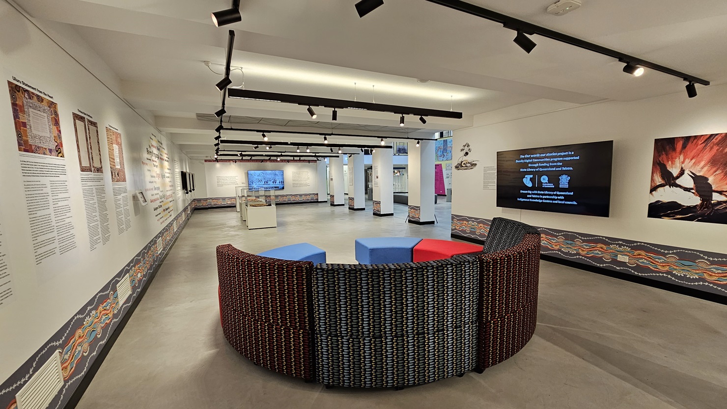The entire wall showcases meticulously painted wall art and art posters. Large flat-screen TVs are mounted on both the right and middle sections of the wall. The ceiling is white, adorned with light fixtures and ceiling art decoration. The floor area is made of polished concrete. In the middle, there is a cushioned seating area arranged in a semi-circle layout. The entire wall showcases meticulously painted wall art and art posters. Large flat-screen TVs are mounted on both the right and middle sections of the wall. The ceiling is white, adorned with light fixtures and ceiling art decoration. The floor area is made of polished concrete. In the middle, there is a cushioned seating area arranged in a semi-circle layout.