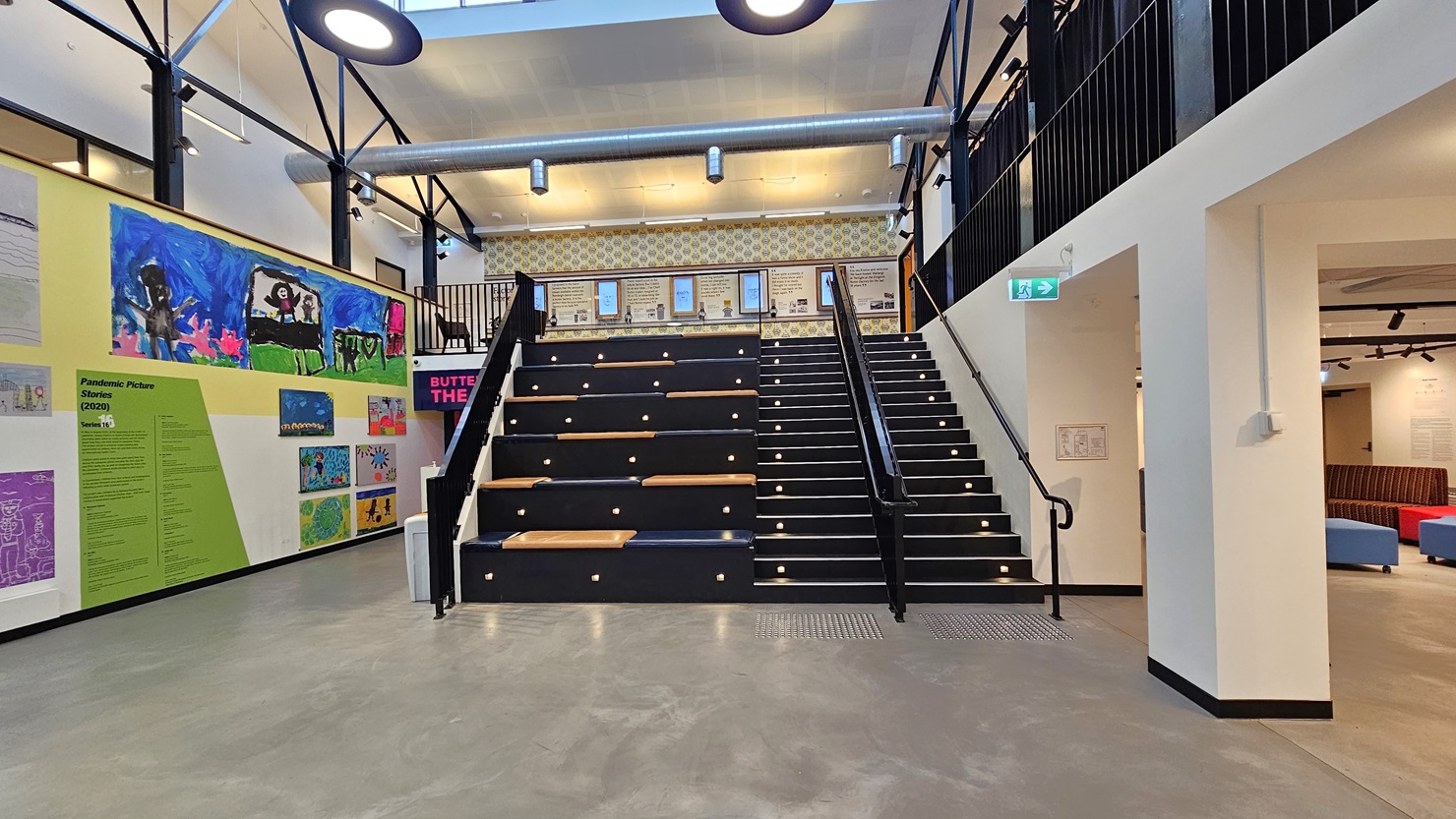 In the centre is a staircase fitted with truncated columns, leading to the mezzanine. To the left, there's a tiered seating area. The left wall is adorned with art images and paintings, with entry to the Butter Theatre on the left. On the right is a seating area with a couch. At the top of the stairs, the mezzanine wall adorned with framed posters is visible. In the centre is a staircase fitted with truncated columns, leading to the mezzanine. To the left, there's a tiered seating area. The left wall is adorned with art images and paintings, with entry to the Butter Theatre on the left. On the right is a seating area with a couch. At the top of the stairs, the mezzanine wall adorned with framed posters is visible.