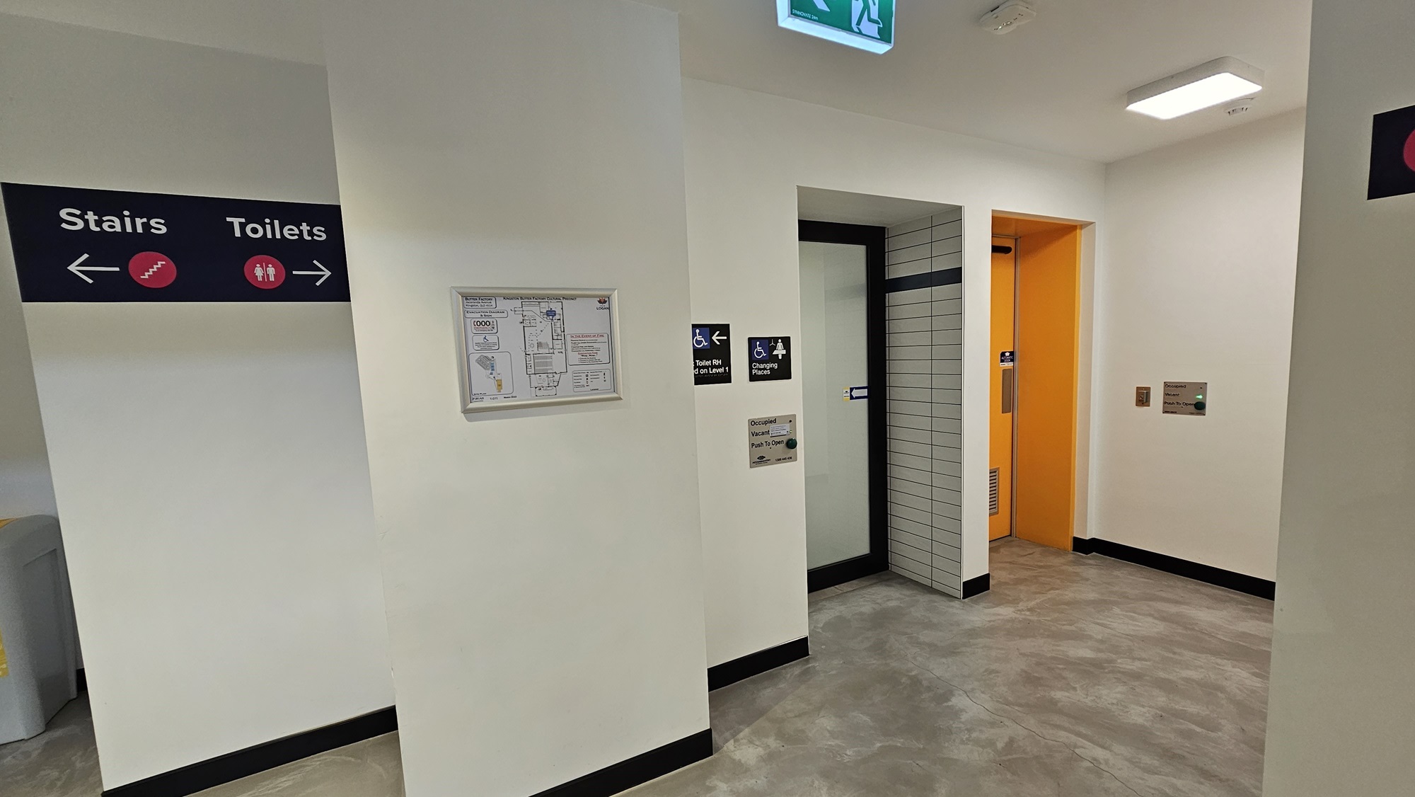 To the left, a wall features directional signage. On the right, is an accessible bathroom and changing place, marked with gender and accessibility signs. The right bathroom has a yellow swing door, while the middle one has a manual white sliding door. A framed poster on the left displays the facility map. The wall is white with tile accents. To the left, a wall features directional signage. On the right, is an accessible bathroom and changing place, marked with gender and accessibility signs. The right bathroom has a yellow swing door, while the middle one has a manual white sliding door. A framed poster on the left displays the facility map. The wall is white with tile accents.