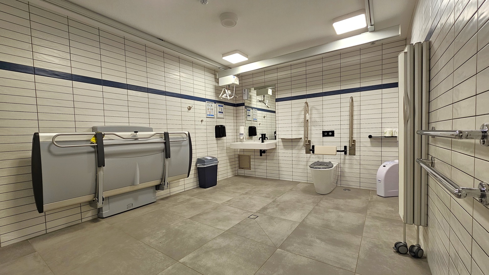 Within the accessible bathroom, an adult-sized change table is affixed to the left wall. A collapsible wall divider stands on the right. In the middle, an ambulant toilet bowl, featuring handrails, is positioned. On the wall, a washbasin, liquid soap dispenser, and tissue dispenser are installed. Tissue rolls are organized on the right, with rubbish bins on both sides. The entire wall is tiled in white, and an additional hand grab is mounted on the right wall. Within the accessible bathroom, an adult-sized change table is affixed to the left wall. A collapsible wall divider stands on the right. In the middle, an ambulant toilet bowl, featuring handrails, is positioned. On the wall, a washbasin, liquid soap dispenser, and tissue dispenser are installed. Tissue rolls are organized on the right, with rubbish bins on both sides. The entire wall is tiled in white, and an additional hand grab is mounted on the right wall.