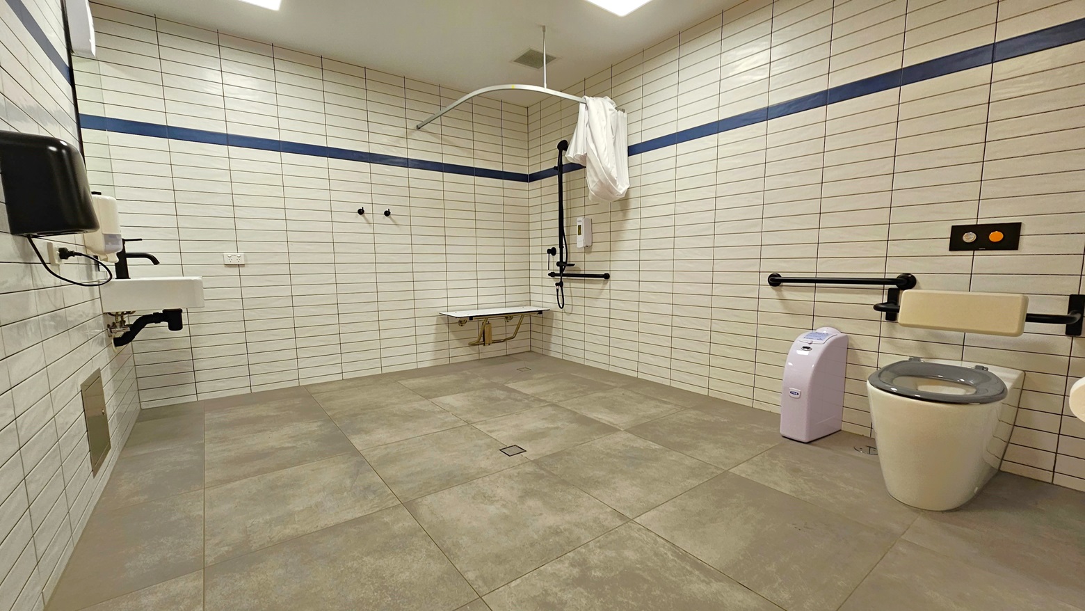 In this spacious bathroom, a shower is situated in the right corner, accompanied by handrails, a bench chair, and a hoist. On the right, a toilet bowl with handrails and a rubbish bin is present. To the left, a washbasin is affixed to the wall, along with a liquid soap dispenser and a hand dryer. The entire area is surrounded by white tiles and white ceilings, while the floor is matte grey. In this spacious bathroom, a shower is situated in the right corner, accompanied by handrails, a bench chair, and a hoist. On the right, a toilet bowl with handrails and a rubbish bin is present. To the left, a washbasin is affixed to the wall, along with a liquid soap dispenser and a hand dryer. The entire area is surrounded by white tiles and white ceilings, while the floor is matte grey.