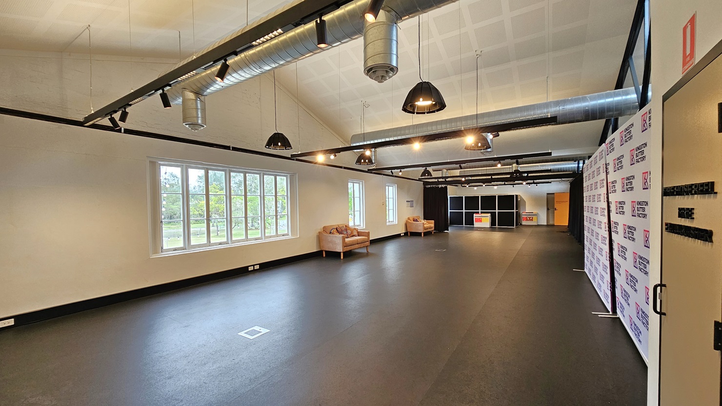 A detailed view of the expansive room reveals cushioned chairs on the left, near large glass windows. On the right, an area is enclosed with navy blue curtains and retractable barriers, featuring a black display rack in the distance. The ceiling is white with exposed air conditioning vents, and light fixtures hang from the white wall. The floor is dark grey. A detailed view of the expansive room reveals cushioned chairs on the left, near large glass windows. On the right, an area is enclosed with navy blue curtains and retractable barriers, featuring a black display rack in the distance. The ceiling is white with exposed air conditioning vents, and light fixtures hang from the white wall. The floor is dark grey.