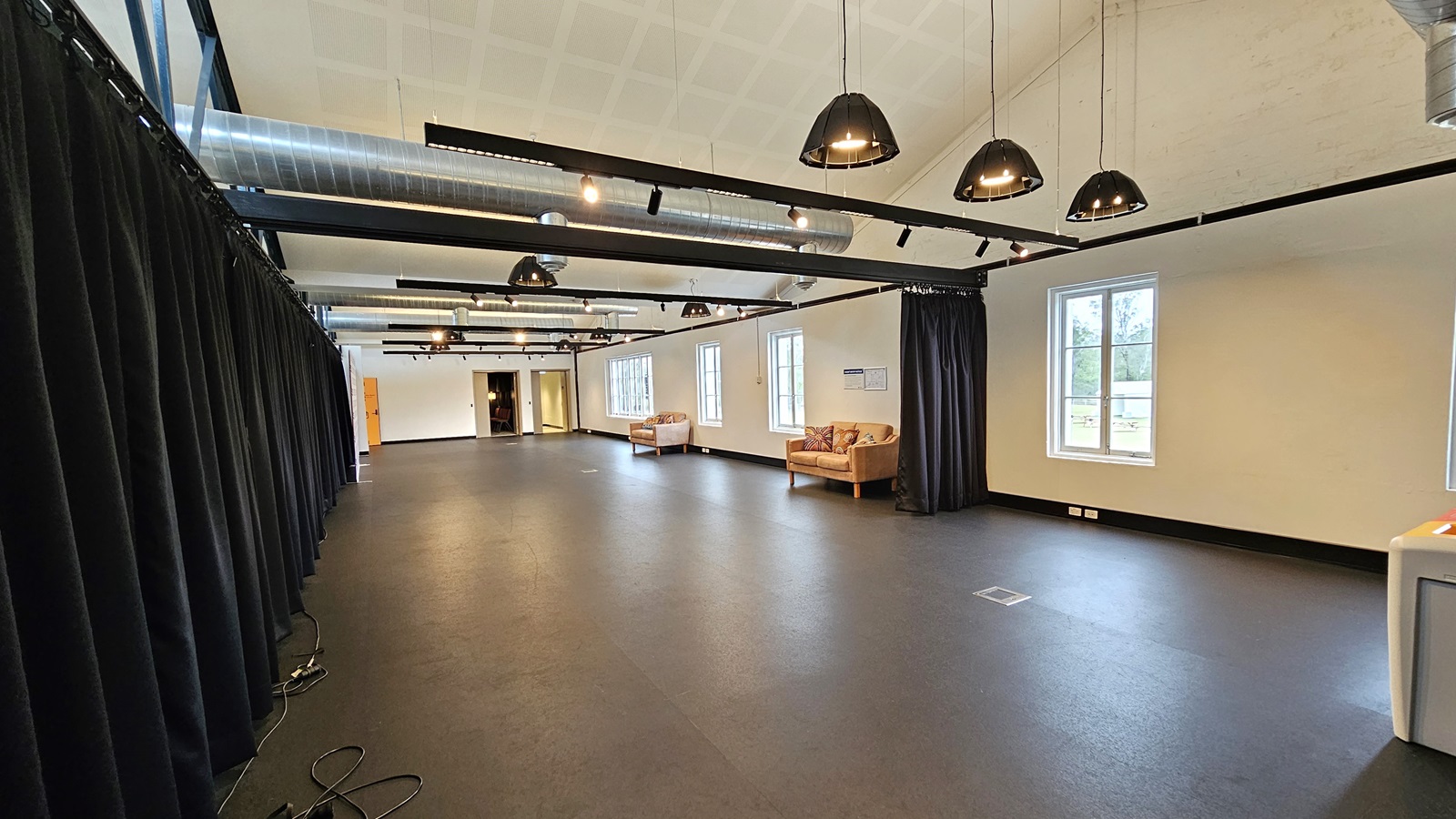 Examining the expansive room, cushioned chairs line the right side near large glass windows. On the left, navy blue curtains and retractable barriers are present. Two doors are situated at the far end. The ceiling is white with exposed air conditioning vents, and light fixtures hang from the white wall. The floor is dark grey, with electrical cords visible on the left floor. Examining the expansive room, cushioned chairs line the right side near large glass windows. On the left, navy blue curtains and retractable barriers are present. Two doors are situated at the far end. The ceiling is white with exposed air conditioning vents, and light fixtures hang from the white wall. The floor is dark grey, with electrical cords visible on the left floor.