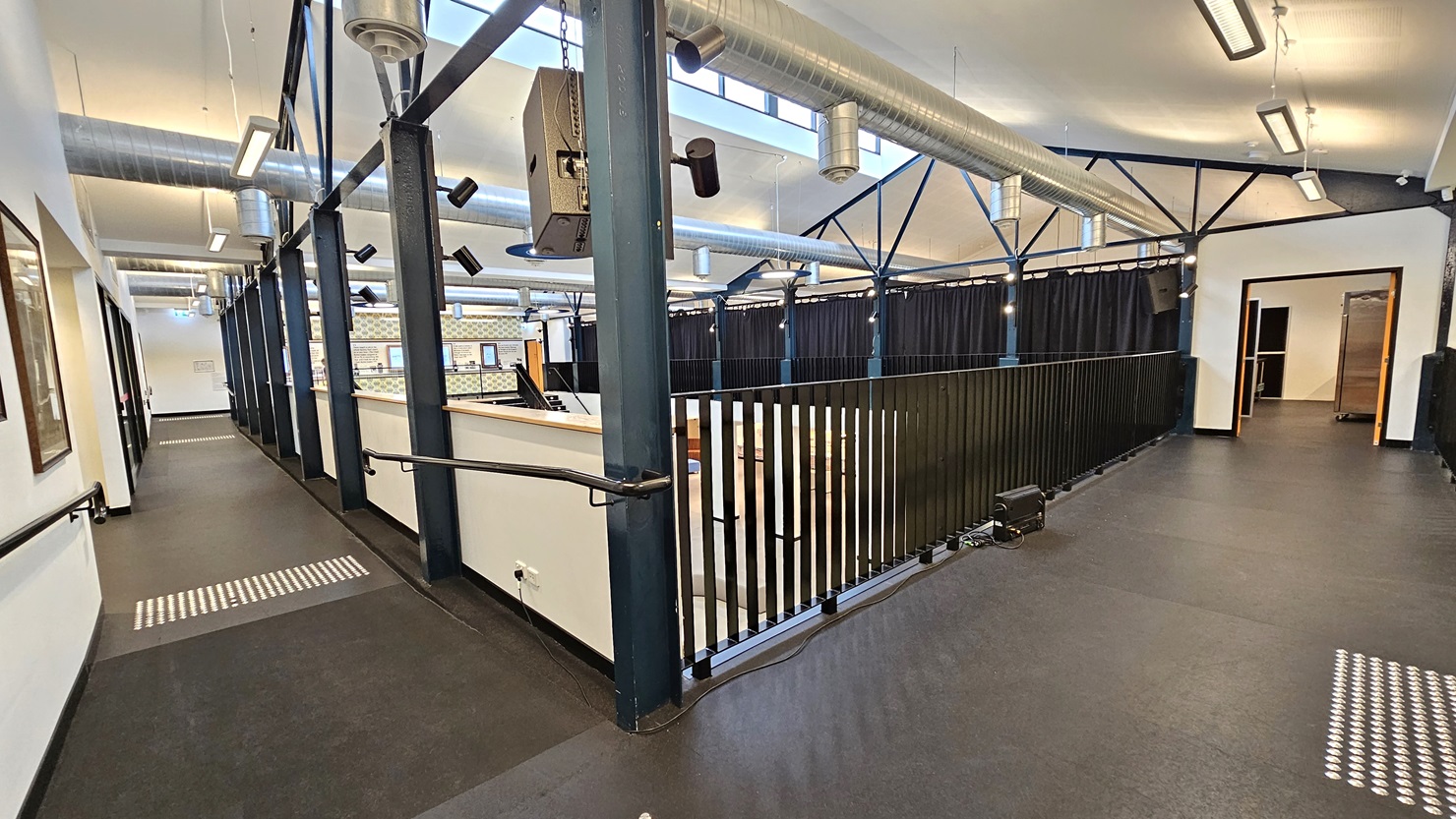To the right is a corridor leading to the other end, secured by concrete railings. Truncated domes are present on the floor. On the left, there's a yellow door, and adjacent to it is a staircase equipped with handrails. Cushioned chairs and a center table with a potted plant are on the right. The floor has grey carpet, and the ceiling adopts a white industrial look with large air conditioning vents and fluorescent lights. To the right is a corridor leading to the other end, secured by concrete railings. Truncated domes are present on the floor. On the left, there's a yellow door, and adjacent to it is a staircase equipped with handrails. Cushioned chairs and a center table with a potted plant are on the right. The floor has grey carpet, and the ceiling adopts a white industrial look with large air conditioning vents and fluorescent lights.