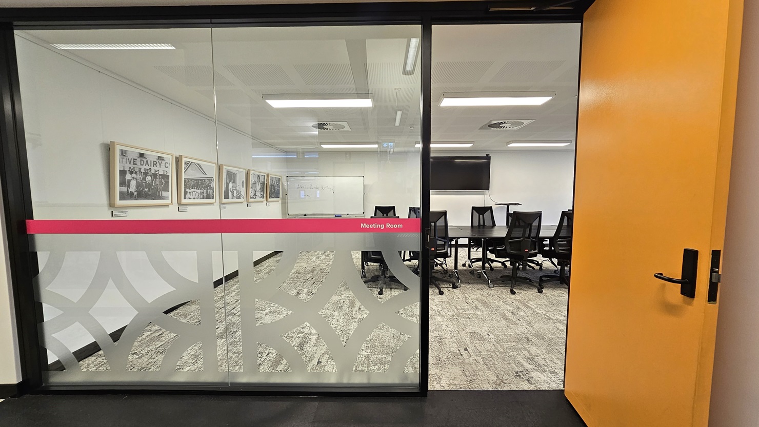 To the left is a glass wall with white and pink print stickers indicating "meeting room." To the right is a yellow wooden door. Inside, a long black conference table is surrounded by black swivel chairs. The left wall features framed posters, and there's a large whiteboard and a flat-screen TV in the middle. The floor has grey carpet, and the ceiling is entirely white with fluorescent lights and centralized air conditioning. To the left is a glass wall with white and pink print stickers indicating "meeting room." To the right is a yellow wooden door. Inside, a long black conference table is surrounded by black swivel chairs. The left wall features framed posters, and there's a large whiteboard and a flat-screen TV in the middle. The floor has grey carpet, and the ceiling is entirely white with fluorescent lights and centralized air conditioning.
