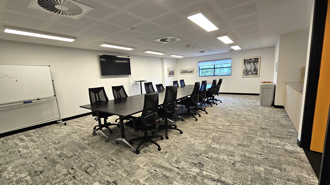 A detailed look at the meeting room reveals a long black conference table in the middle, surrounded by black swivel chairs. The central wall has a large glass window and framed posters. On the left wall, there's a large whiteboard and a flat-screen TV. The floor is covered with grey tiles, and the ceiling is entirely white, featuring fluorescent lights and centralized air conditioning. A detailed look at the meeting room reveals a long black conference table in the middle, surrounded by black swivel chairs. The central wall has a large glass window and framed posters. On the left wall, there's a large whiteboard and a flat-screen TV. The floor is covered with grey tiles, and the ceiling is entirely white, featuring fluorescent lights and centralized air conditioning.
