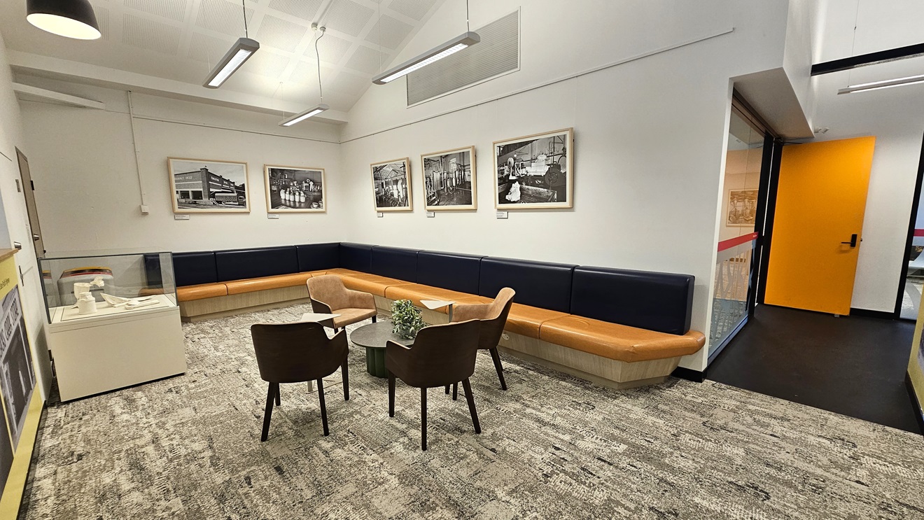 The seating area comprises an L-shaped black and brown couch with a central table surrounded by seating, adorned with a potted plant. The wall is decorated with glass-framed posters, and on the left is a table with glass panels. The floor is covered with grey carpet, and the ceiling is entirely white, featuring fluorescent lights and centralized air conditioning. On the right, there is a glass wall and a yellow door. The seating area comprises an L-shaped black and brown couch with a central table surrounded by seating, adorned with a potted plant. The wall is decorated with glass-framed posters, and on the left is a table with glass panels. The floor is covered with grey carpet, and the ceiling is entirely white, featuring fluorescent lights and centralized air conditioning. On the right, there is a glass wall and a yellow door.