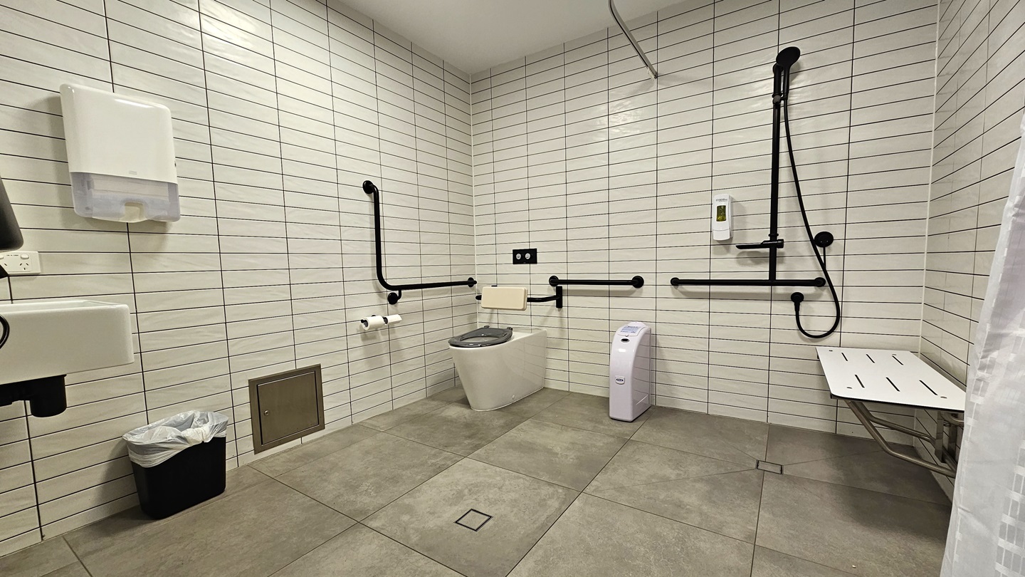 The spacious bathroom includes a corner toilet with handrails and a mounted black shower. A tissue dispenser is located beside the toilet. On the right, a wall-mounted bench chair and a white curtain are positioned. To the left, a washbasin and another tissue dispenser are available, with a rubbish bin underneath. The walls are covered in white tiles, and the ceiling is white. The floor is tiled in grey. The spacious bathroom includes a corner toilet with handrails and a mounted black shower. A tissue dispenser is located beside the toilet. On the right, a wall-mounted bench chair and a white curtain are positioned. To the left, a washbasin and another tissue dispenser are available, with a rubbish bin underneath. The walls are covered in white tiles, and the ceiling is white. The floor is tiled in grey.