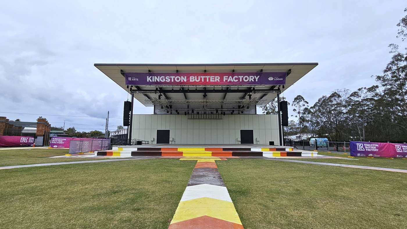 A concrete pathway runs across the grassy area toward the stage. The stage has colorful steps and white wall tiles. A gradient pink and blue banner displaying the facility's name hangs near the ceiling. On the left, the Butter Factory building is present with a red brick facade and gradient banners. Picnic tables are positioned in the grassy area. A concrete pathway runs across the grassy area toward the stage. The stage has colorful steps and white wall tiles. A gradient pink and blue banner displaying the facility's name hangs near the ceiling. On the left, the Butter Factory building is present with a red brick facade and gradient banners. Picnic tables are positioned in the grassy area.