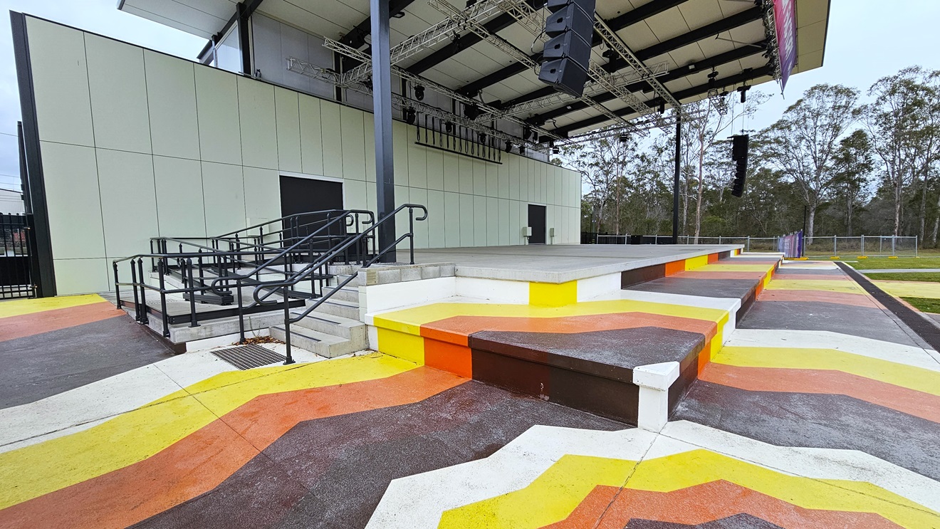 The stage steps are adorned with colourful lines in yellow, orange, brown, and white. To the left, there are additional entries, including stairs and a ramp with handrails and truncated domes. The stage exhibits white walls and an industrial ceiling with exposed beams. Large audio speakers are suspended on both sides of the stage. The surroundings include trees and grass. The stage steps are adorned with colourful lines in yellow, orange, brown, and white. To the left, there are additional entries, including stairs and a ramp with handrails and truncated domes. The stage exhibits white walls and an industrial ceiling with exposed beams. Large audio speakers are suspended on both sides of the stage. The surroundings include trees and grass.