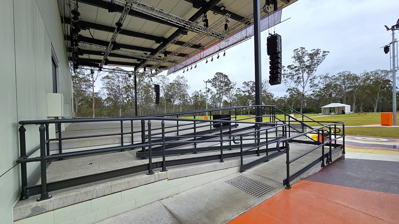 A wheelchair-accessible entry is situated on the left of the stage, featuring a ramp with handrails and truncated domes on the floor. The stage displays white walls and an industrial ceiling with exposed beams. Large audio speakers are suspended on both sides of the stage. Colourfully painted steps adorn the front of the stage, and a lamp post stands in the grassy area. A wheelchair-accessible entry is situated on the left of the stage, featuring a ramp with handrails and truncated domes on the floor. The stage displays white walls and an industrial ceiling with exposed beams. Large audio speakers are suspended on both sides of the stage. Colourfully painted steps adorn the front of the stage, and a lamp post stands in the grassy area.