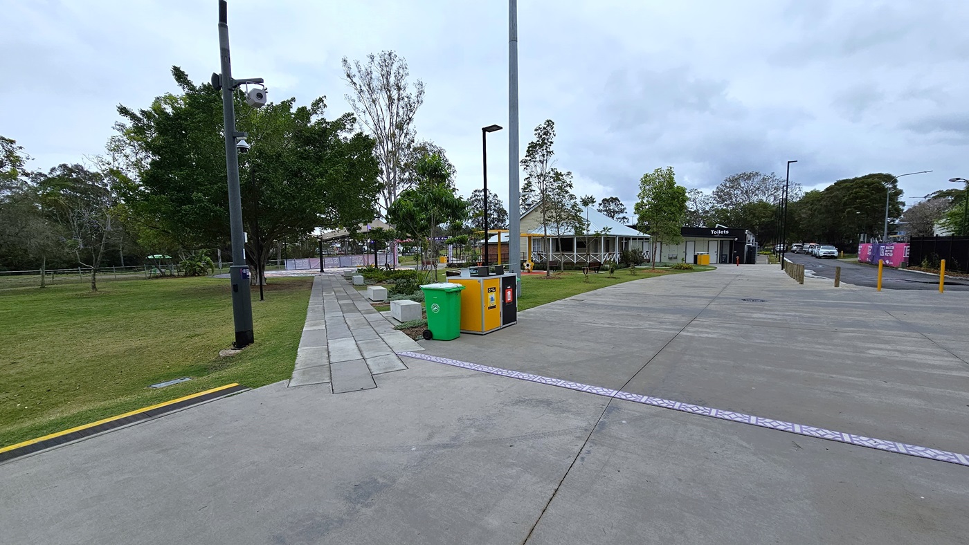 A concrete path extends to the grassy area, bordered by landscaping and trees on the right. Additional trees line the left side, while tall light posts and building structures stand in the middle. On the right, a concrete road leads to the car park, bordered by yellow truncated domes. A concrete path extends to the grassy area, bordered by landscaping and trees on the right. Additional trees line the left side, while tall light posts and building structures stand in the middle. On the right, a concrete road leads to the car park, bordered by yellow truncated domes.
