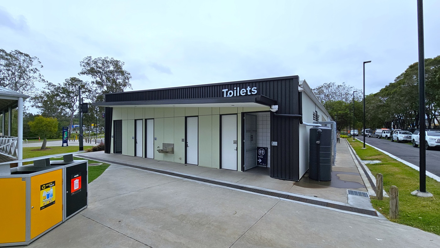A toilet block, featuring 6 white doors marked for gender access, is situated near the road. The building has corrugated design black walls. On the left, yellow and black rubbish bins are marked for separating recyclables. A section of a building structure is on the left, while to the right, a road accommodates a few parked cars. A toilet block, featuring 6 white doors marked for gender access, is situated near the road. The building has corrugated design black walls. On the left, yellow and black rubbish bins are marked for separating recyclables. A section of a building structure is on the left, while to the right, a road accommodates a few parked cars.