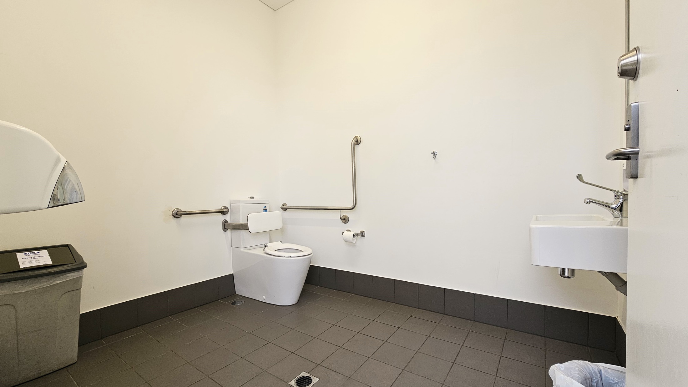 Inside the toilet, a toilet bowl equipped with handrails is positioned in the corner along with a tissue dispenser. To the right, a washbasin is mounted, and a rubbish bin is placed underneath. On the left, a tissue dispenser is mounted to the wall with a rubbish bin underneath. The wall is white, and the floor has grey tiles. Inside the toilet, a toilet bowl equipped with handrails is positioned in the corner along with a tissue dispenser. To the right, a washbasin is mounted, and a rubbish bin is placed underneath. On the left, a tissue dispenser is mounted to the wall with a rubbish bin underneath. The wall is white, and the floor has grey tiles.
