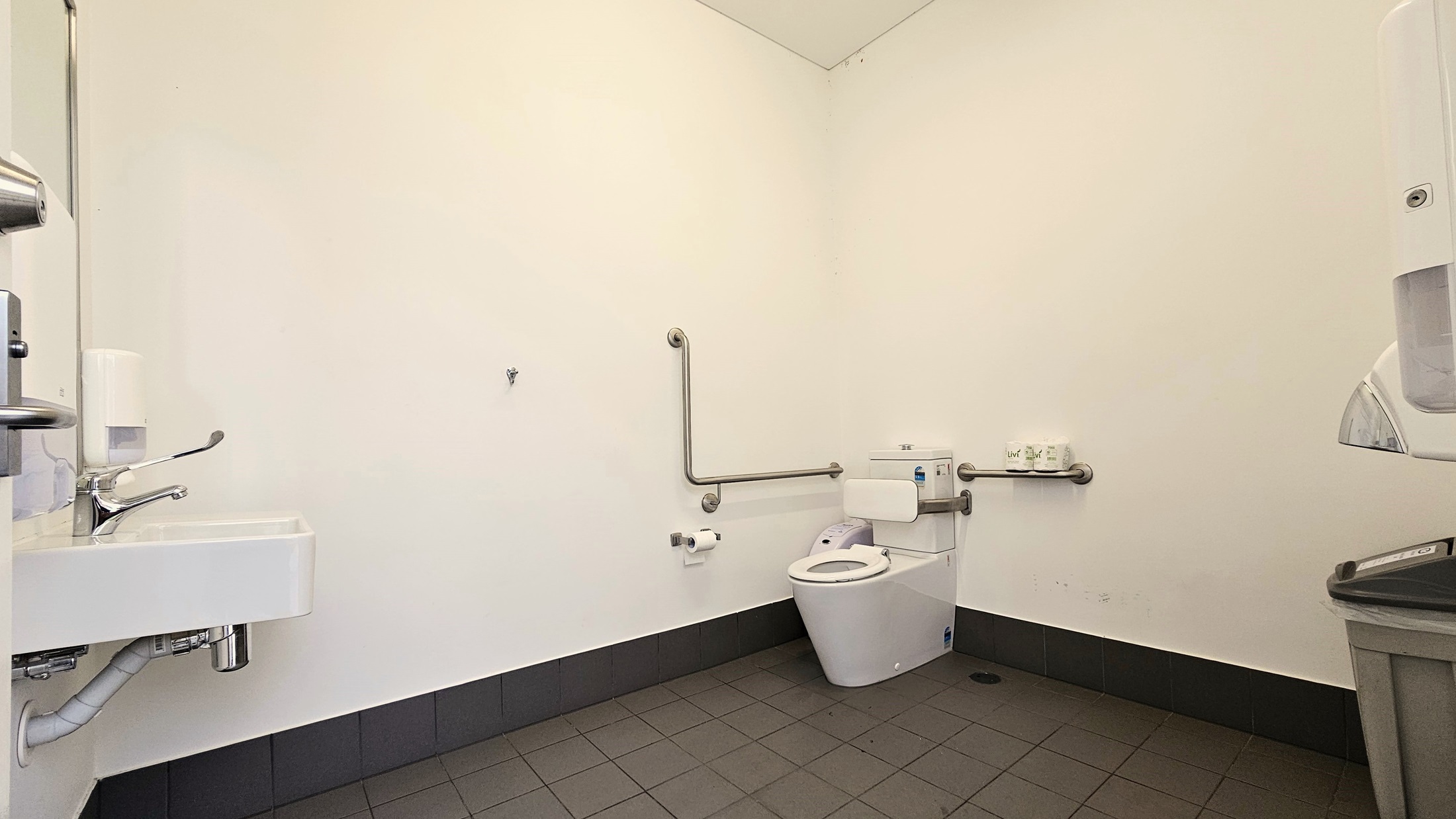 Inside the toilet, a toilet bowl equipped with handrails is positioned in the corner, accompanied by a tissue dispenser. On the left, a washbasin with a liquid soap dispenser is mounted on the wall, and a rubbish bin is placed underneath. On the right, a tissue dispenser is mounted to the wall with a rubbish bin underneath. The wall is white, and the floor has grey tiles. Inside the toilet, a toilet bowl equipped with handrails is positioned in the corner, accompanied by a tissue dispenser. On the left, a washbasin with a liquid soap dispenser is mounted on the wall, and a rubbish bin is placed underneath. On the right, a tissue dispenser is mounted to the wall with a rubbish bin underneath. The wall is white, and the floor has grey tiles.