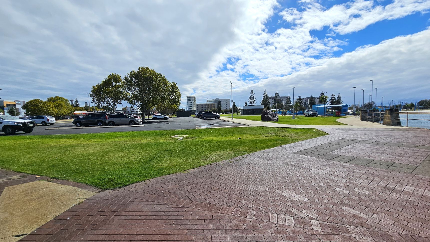 A paved area with a mix of red and grey bricks is in the foreground, leading to a grassy section. To the left, several cars are parked near a row of trees. In the background, there are buildings and more parked cars. On the right, a pathway curves towards a waterfront area with a low stone wall and a body of water. The sky is partly cloudy with patches of blue visible. A paved area with a mix of red and grey bricks is in the foreground, leading to a grassy section. To the left, several cars are parked near a row of trees. In the background, there are buildings and more parked cars. On the right, a pathway curves towards a waterfront area with a low stone wall and a body of water. The sky is partly cloudy with patches of blue visible.