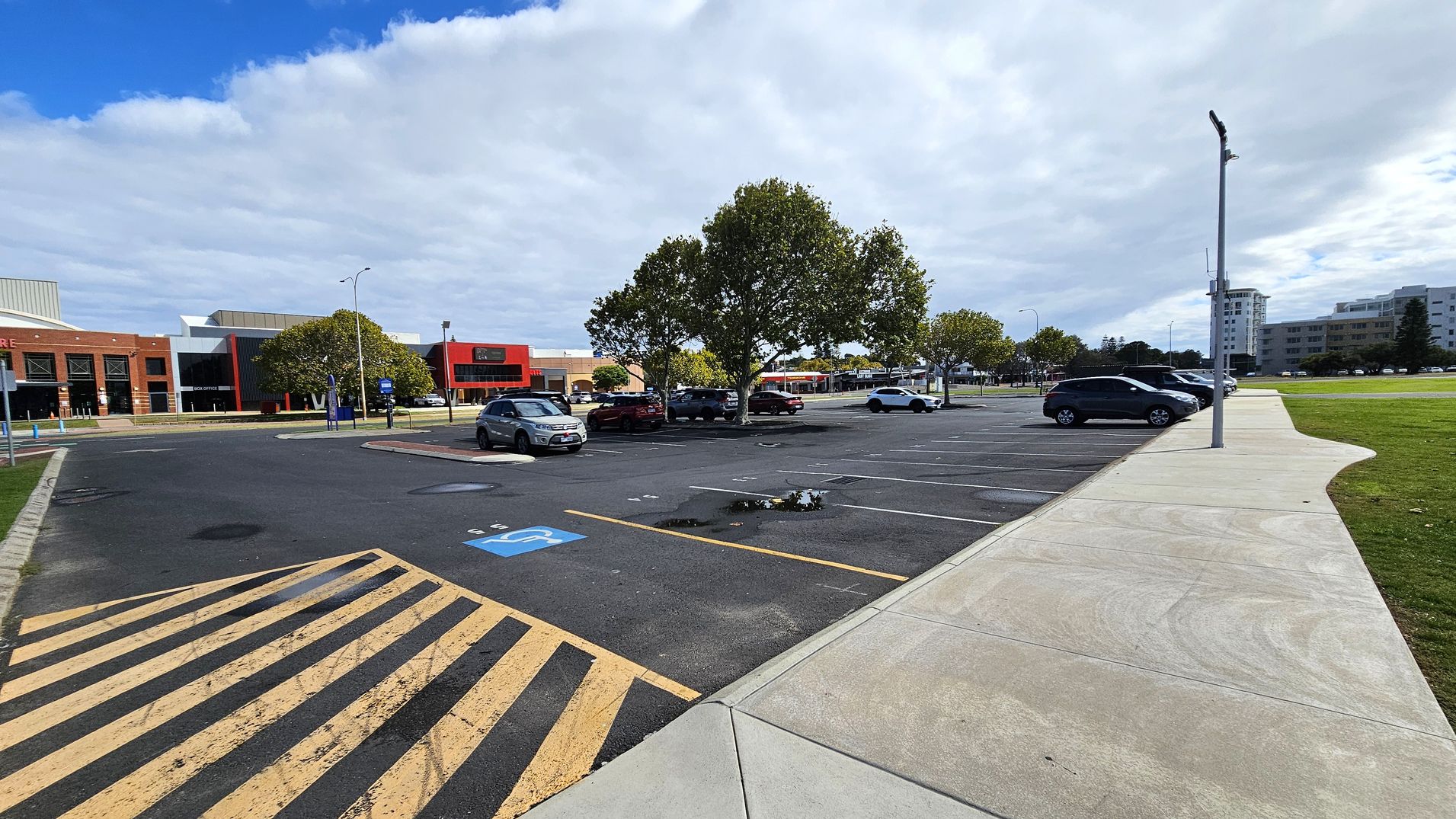 A car park with several vehicles is visible, with a row of trees in the centre. On the left, there is a brick building with a red and black facade. The foreground shows a pedestrian crossing with yellow and black stripes and a disabled parking space. On the right, a concrete footpath runs alongside the parking spaces, leading to a grassy area. In the background, there are more buildings, including a tall white one. A streetlamp is positioned near the footpath. A car park with several vehicles is visible, with a row of trees in the centre. On the left, there is a brick building with a red and black facade. The foreground shows a pedestrian crossing with yellow and black stripes and a disabled parking space. On the right, a concrete footpath runs alongside the parking spaces, leading to a grassy area. In the background, there are more buildings, including a tall white one. A streetlamp is positioned near the footpath.
