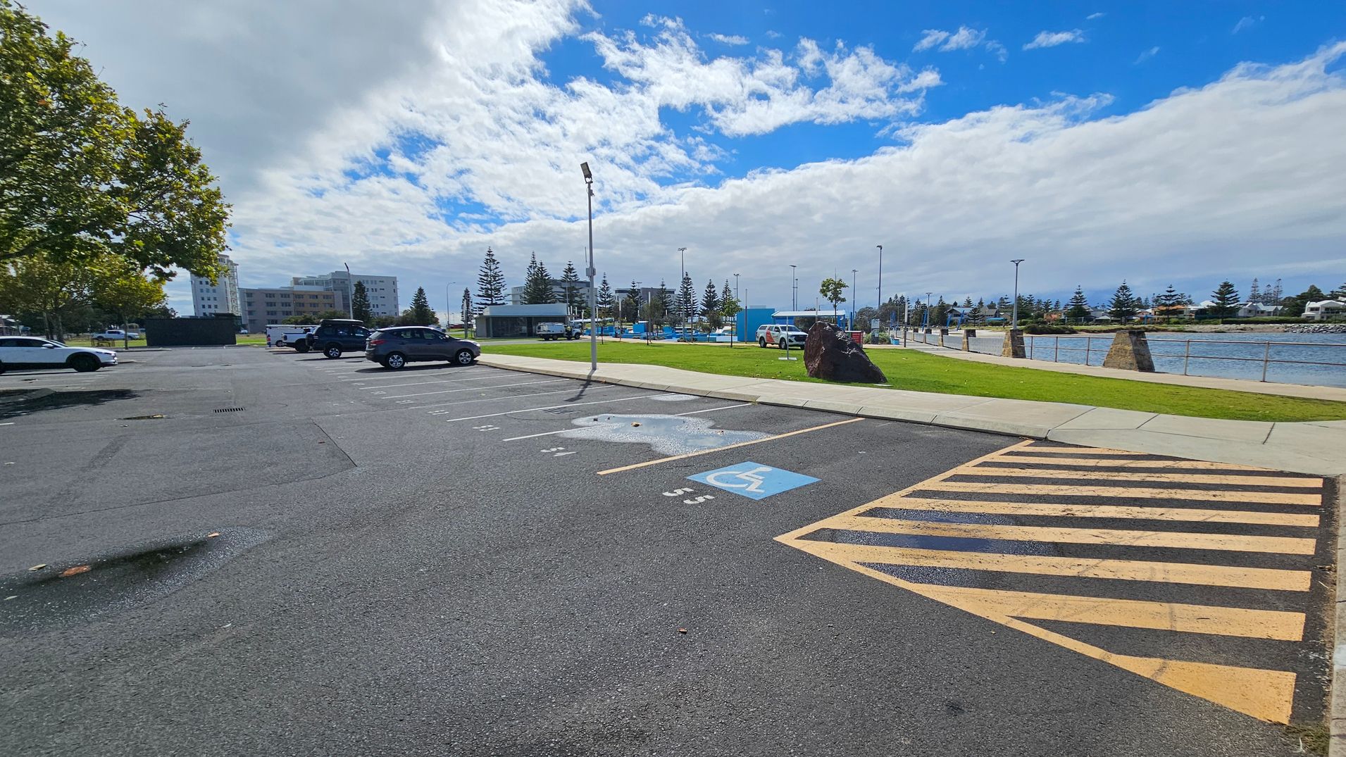 A car park with marked spaces, including a designated disabled parking spot with a blue symbol, is in the foreground. To the right, a yellow-striped area is visible. In the background, there is a grassy area with a large rock, a few trees, and a body of water with a railing along the edge. Several buildings and parked cars are on the left, with a tree providing some shade. The sky is partly cloudy. A car park with marked spaces, including a designated disabled parking spot with a blue symbol, is in the foreground. To the right, a yellow-striped area is visible. In the background, there is a grassy area with a large rock, a few trees, and a body of water with a railing along the edge. Several buildings and parked cars are on the left, with a tree providing some shade. The sky is partly cloudy.