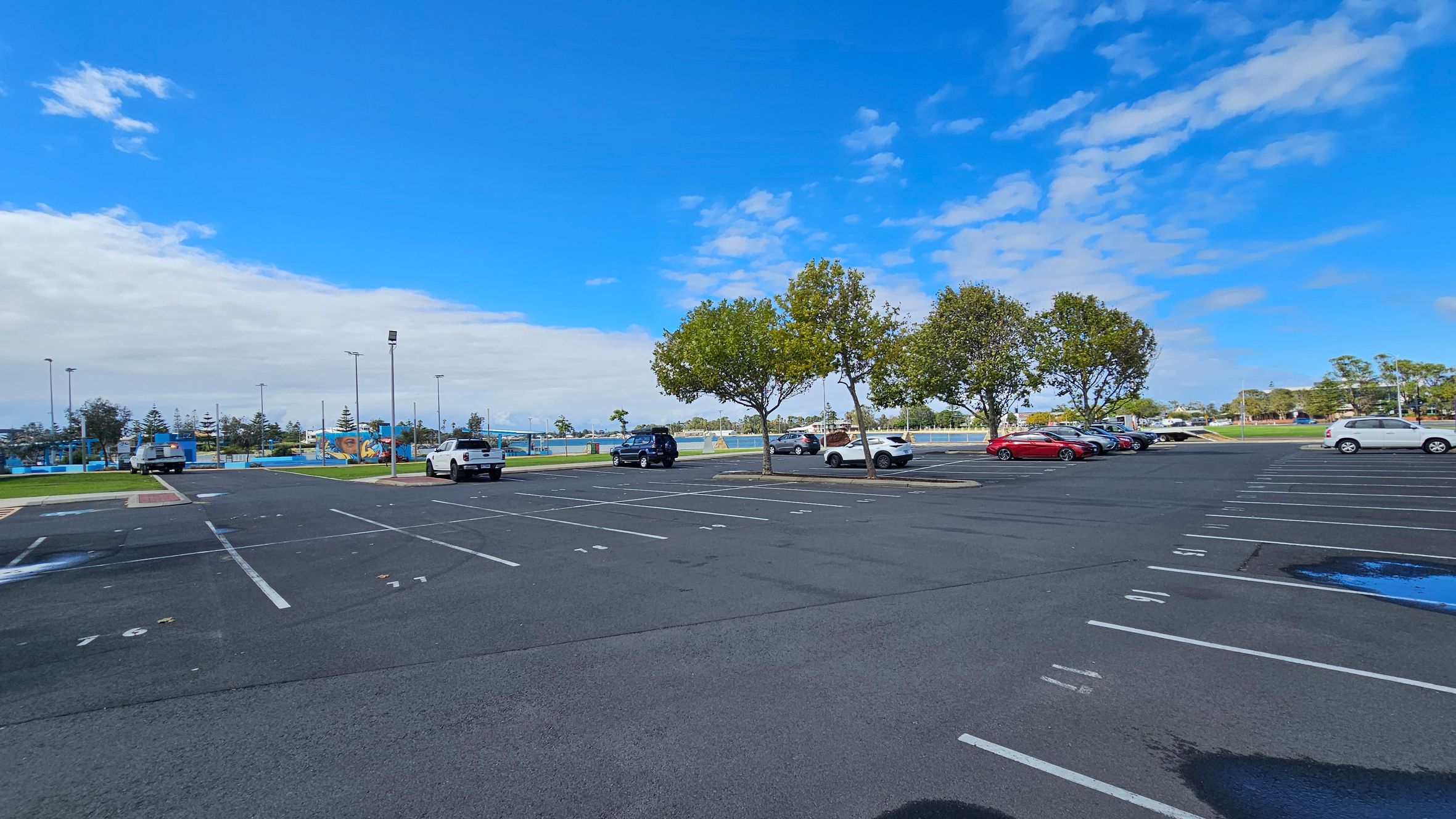 An expansive car park with marked parking spaces, some occupied by vehicles. On the right, several cars are parked near a row of trees. In the middle, a few more cars are scattered across the lot. The left side shows a grassy area with a few trees and a distant view of a sports field or park. The ground is asphalt with visible parking lines, and there are a few puddles. The background features more trees and some structures, possibly part of a recreational area. An expansive car park with marked parking spaces, some occupied by vehicles. On the right, several cars are parked near a row of trees. In the middle, a few more cars are scattered across the lot. The left side shows a grassy area with a few trees and a distant view of a sports field or park. The ground is asphalt with visible parking lines, and there are a few puddles. The background features more trees and some structures, possibly part of a recreational area.