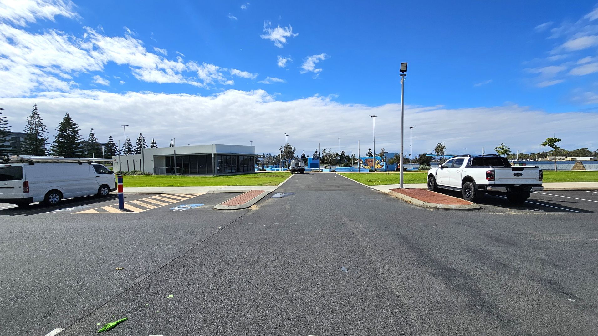 A car park with a white van on the left and a white pickup truck on the right. In the centre, a road leads to a building with large windows and a flat roof. The background features a grassy area with scattered trees and a playground. A tall streetlight is on the right side of the road. The sky is partly cloudy. A car park with a white van on the left and a white pickup truck on the right. In the centre, a road leads to a building with large windows and a flat roof. The background features a grassy area with scattered trees and a playground. A tall streetlight is on the right side of the road. The sky is partly cloudy.