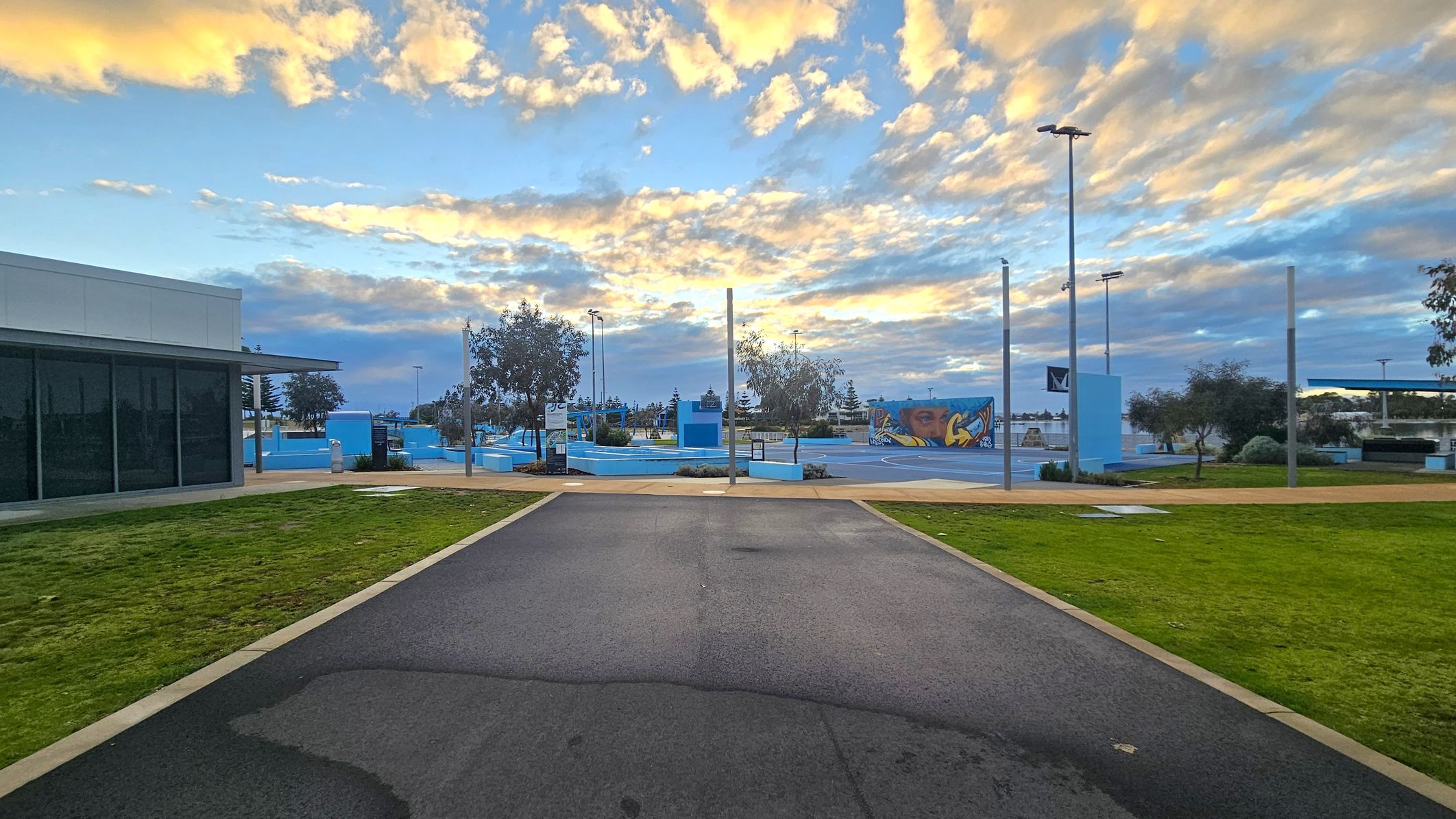 A paved path leads towards a skate park in the centre, with blue ramps and a colourful mural. On the left, a building with large dark windows and a flat roof. On the right, green grass and scattered trees. Tall light poles are visible throughout the area. The sky is filled with clouds tinged with yellow and orange hues. A paved path leads towards a skate park in the centre, with blue ramps and a colourful mural. On the left, a building with large dark windows and a flat roof. On the right, green grass and scattered trees. Tall light poles are visible throughout the area. The sky is filled with clouds tinged with yellow and orange hues.
