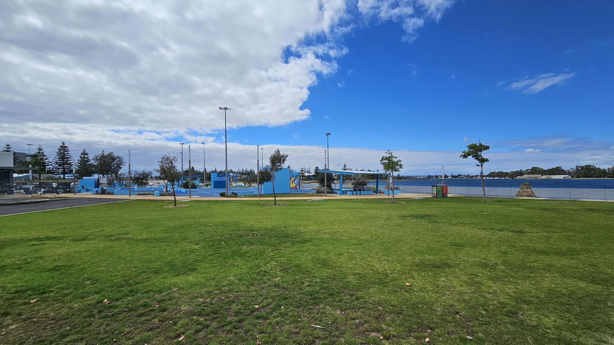 A grassy park area with patches of green and brown in the foreground. In the middle, there are several small trees and a pathway leading to a blue-painted recreational area with structures and poles. To the right, a body of water is visible, bordered by a fence. The background features more trees and a partly cloudy sky. A grassy park area with patches of green and brown in the foreground. In the middle, there are several small trees and a pathway leading to a blue-painted recreational area with structures and poles. To the right, a body of water is visible, bordered by a fence. The background features more trees and a partly cloudy sky.