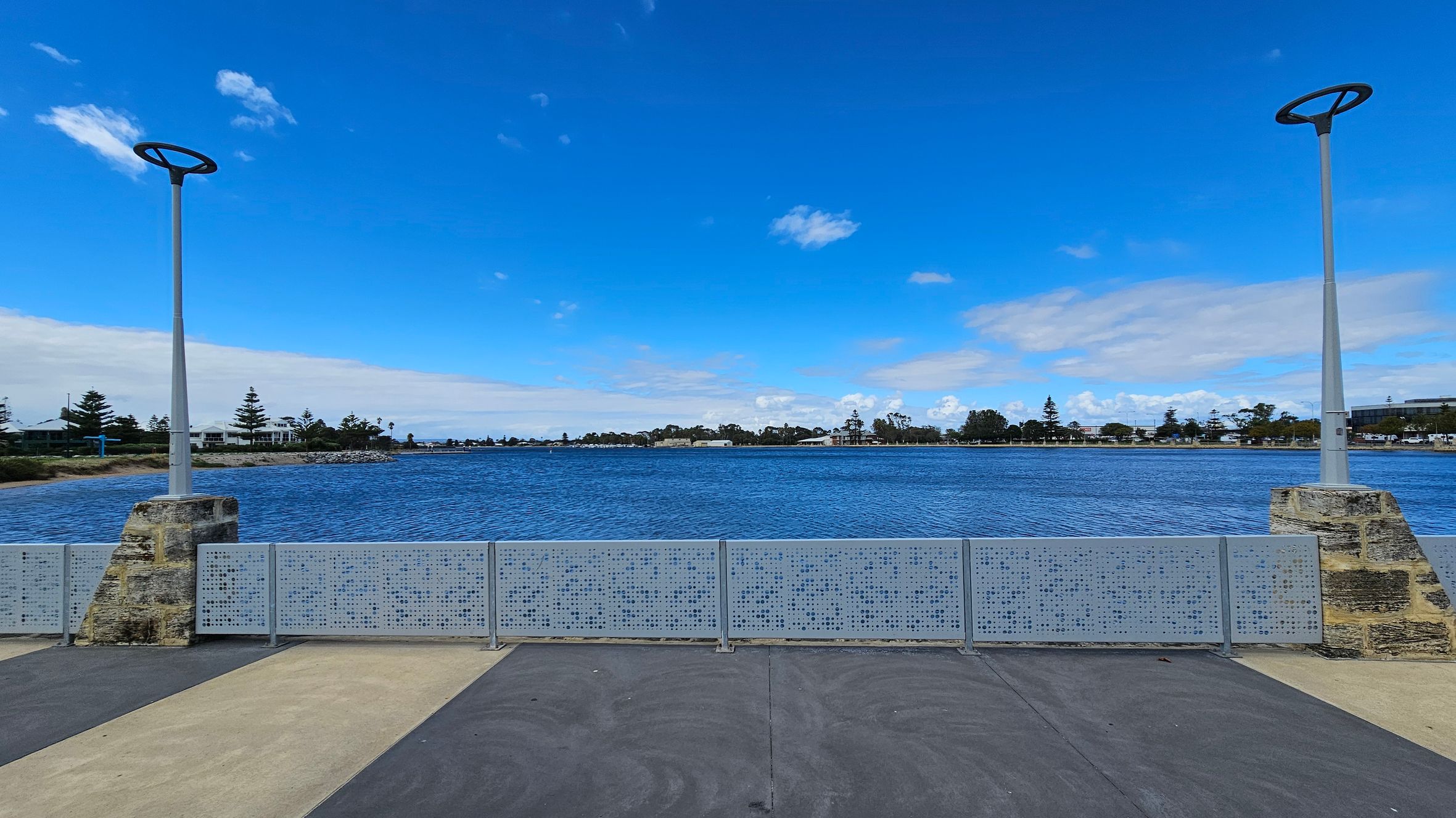 A waterfront scene with a wide expanse of blue water in the middle. In the foreground, there is a paved area with alternating light and dark sections. Two modern street lamps with circular tops are positioned on stone bases, one on the left and one on the right. A metal railing with a perforated design runs across the middle. In the background, there are trees and buildings along the shoreline under a clear blue sky. A waterfront scene with a wide expanse of blue water in the middle. In the foreground, there is a paved area with alternating light and dark sections. Two modern street lamps with circular tops are positioned on stone bases, one on the left and one on the right. A metal railing with a perforated design runs across the middle. In the background, there are trees and buildings along the shoreline under a clear blue sky.