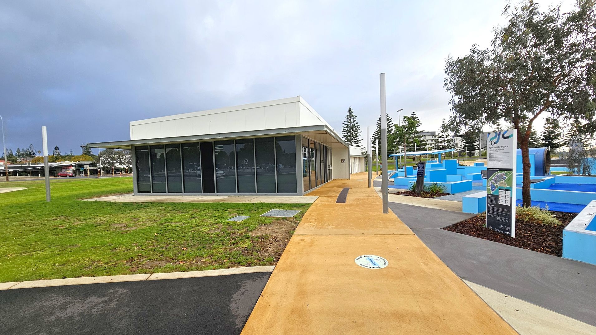 A modern, single-storey building with large glass windows on the left, surrounded by a grassy area. A paved path leads to the building, with a circular sign on the ground. On the right, there are blue geometric structures and a sign with information. A tree is nearby, and several tall poles are scattered around. In the background, there are more trees and a few buildings. The sky is overcast. A modern, single-storey building with large glass windows on the left, surrounded by a grassy area. A paved path leads to the building, with a circular sign on the ground. On the right, there are blue geometric structures and a sign with information. A tree is nearby, and several tall poles are scattered around. In the background, there are more trees and a few buildings. The sky is overcast.
