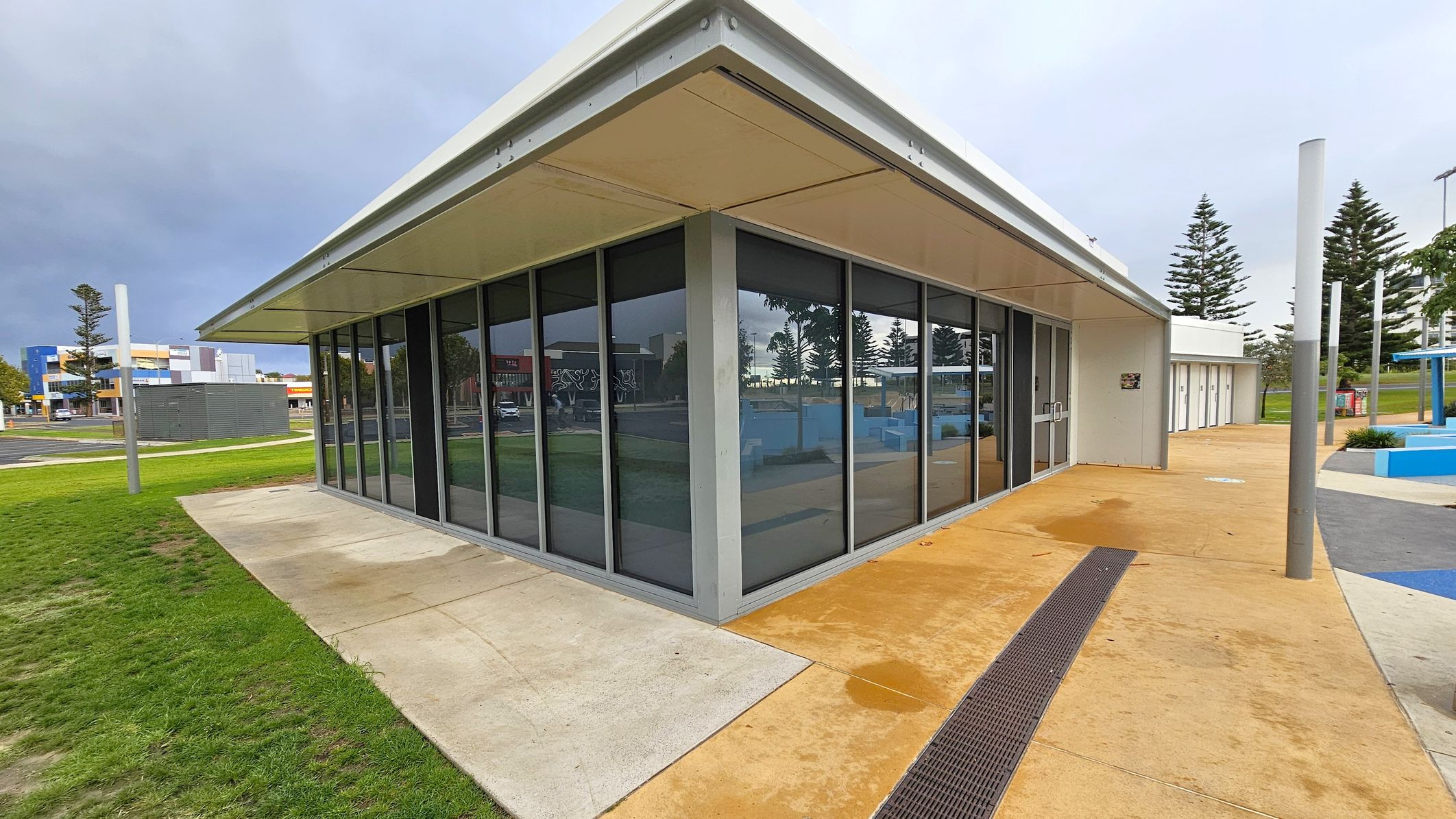 A modern building with large glass windows on the left, set on a concrete path that transitions into grass. The right side features a continuation of the building with a beige walkway and a series of doors. Tall, slender poles are positioned along the path, and several pine trees are visible in the background. The ceiling is flat and white, matching the building's exterior. A modern building with large glass windows on the left, set on a concrete path that transitions into grass. The right side features a continuation of the building with a beige walkway and a series of doors. Tall, slender poles are positioned along the path, and several pine trees are visible in the background. The ceiling is flat and white, matching the building's exterior.
