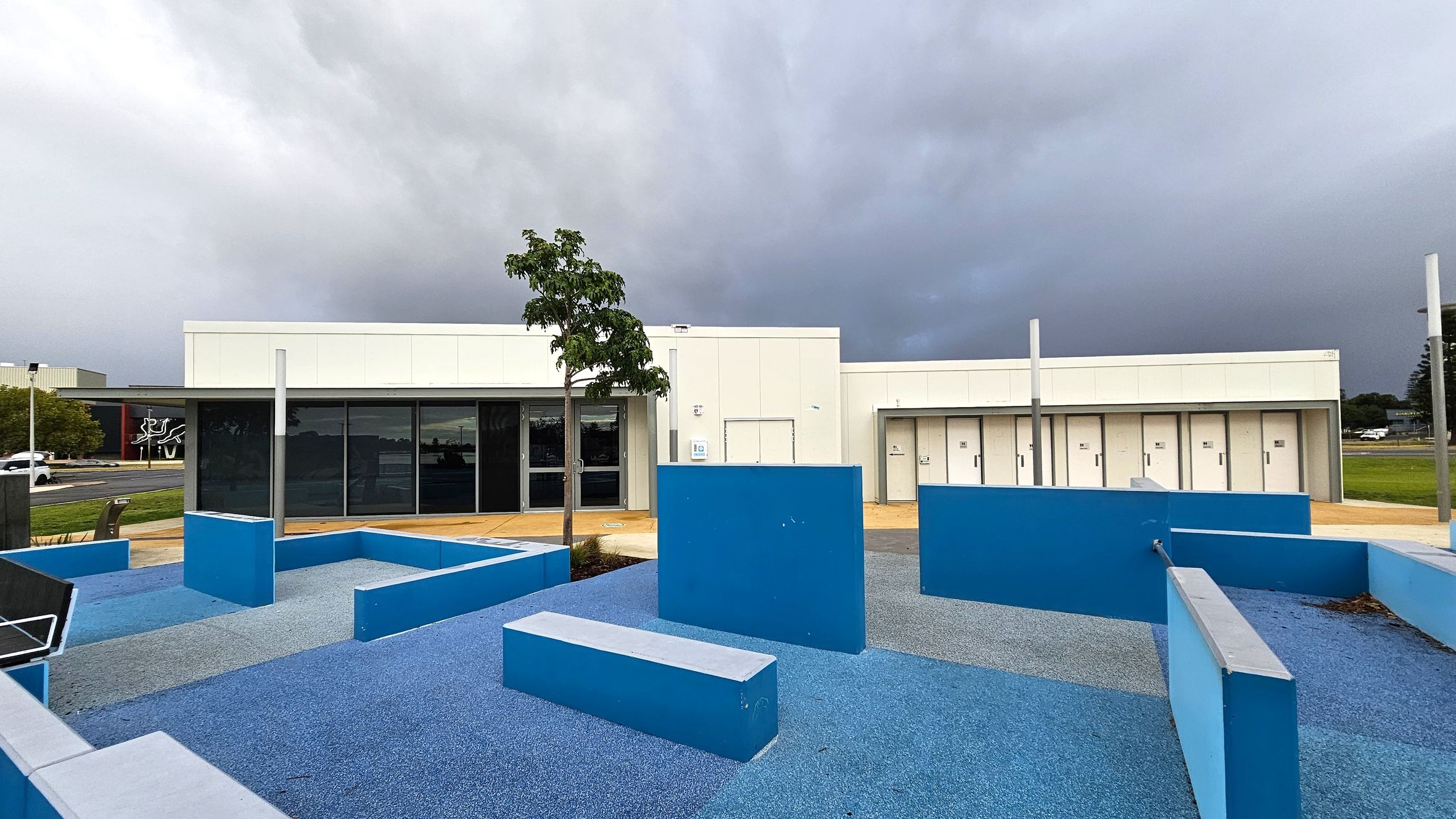 A modern building with a flat roof and white exterior is in the background. In front, there is a playground area with blue rubber flooring and various blue geometric structures, including benches and walls. A small tree is planted near the centre. To the left, a road with a few cars and a building with a mural can be seen. The sky is overcast, creating a grey backdrop. A modern building with a flat roof and white exterior is in the background. In front, there is a playground area with blue rubber flooring and various blue geometric structures, including benches and walls. A small tree is planted near the centre. To the left, a road with a few cars and a building with a mural can be seen. The sky is overcast, creating a grey backdrop.