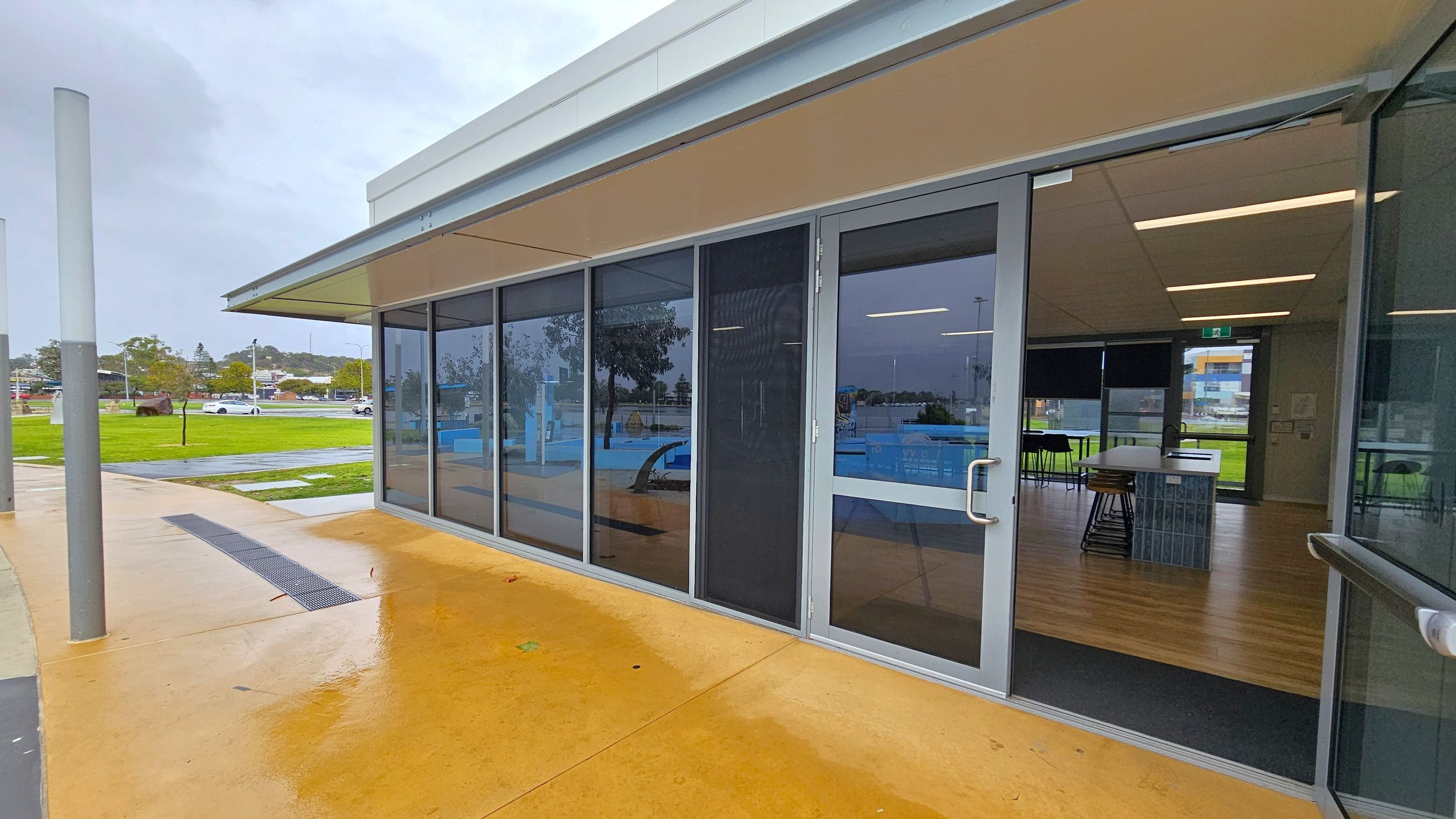 A modern building with large glass windows and doors on the right, reflecting the surroundings. Inside, there are tables and chairs on a wooden floor. The ceiling has recessed lighting. Outside, the ground is wet, suggesting recent rain, with a yellowish concrete surface. In the background, there is a grassy area with trees and parked cars. A tall, cylindrical post is visible on the left. A modern building with large glass windows and doors on the right, reflecting the surroundings. Inside, there are tables and chairs on a wooden floor. The ceiling has recessed lighting. Outside, the ground is wet, suggesting recent rain, with a yellowish concrete surface. In the background, there is a grassy area with trees and parked cars. A tall, cylindrical post is visible on the left.