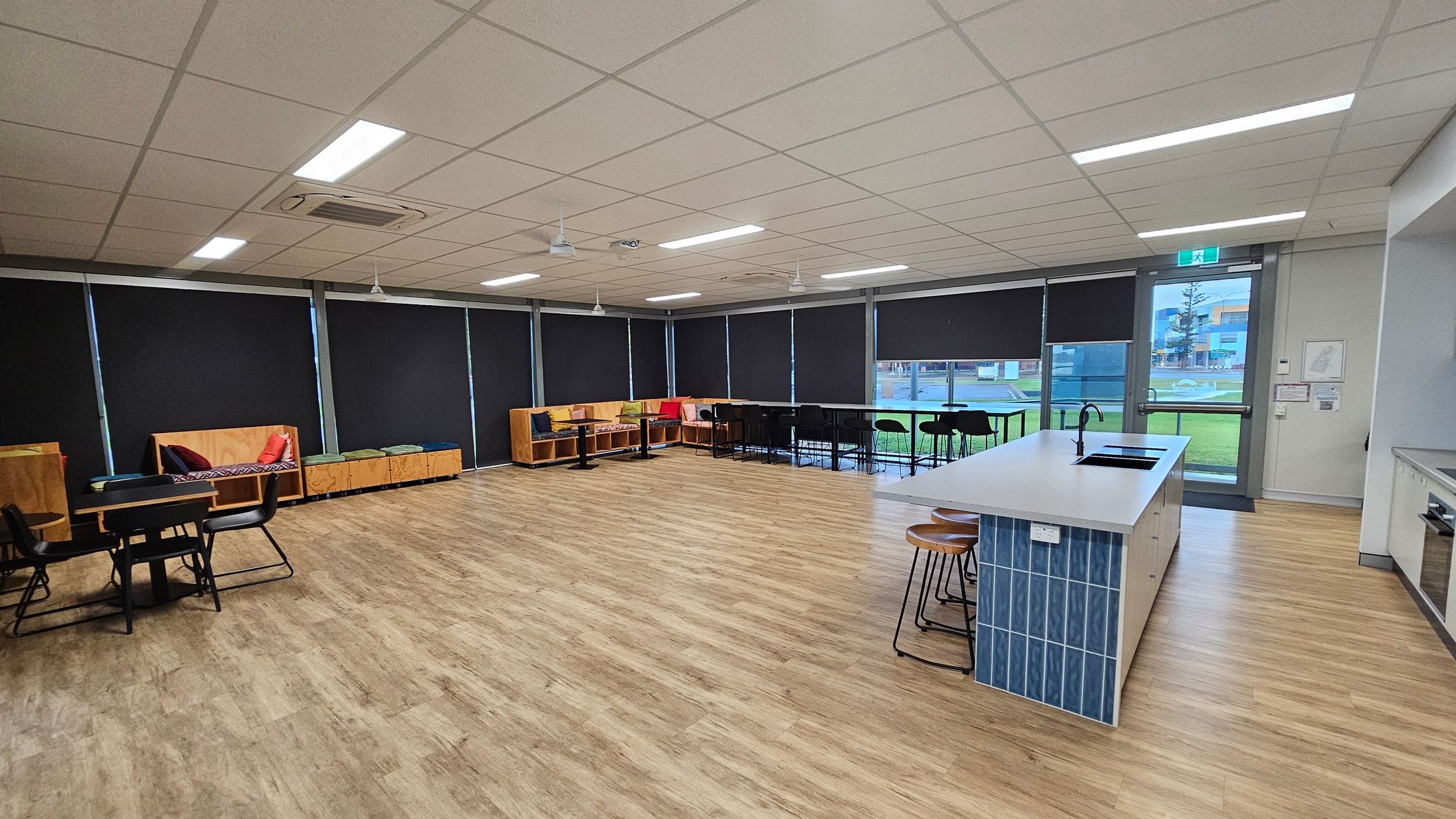A spacious room with a light wooden floor and a white tiled ceiling. On the left, there are tables with black chairs and wooden benches with colourful cushions. The right side features a kitchen island with blue tiles and wooden stools. Large windows with black blinds line the back wall, and a glass door leads outside. The room is well-lit with fluorescent lights. A spacious room with a light wooden floor and a white tiled ceiling. On the left, there are tables with black chairs and wooden benches with colourful cushions. The right side features a kitchen island with blue tiles and wooden stools. Large windows with black blinds line the back wall, and a glass door leads outside. The room is well-lit with fluorescent lights.